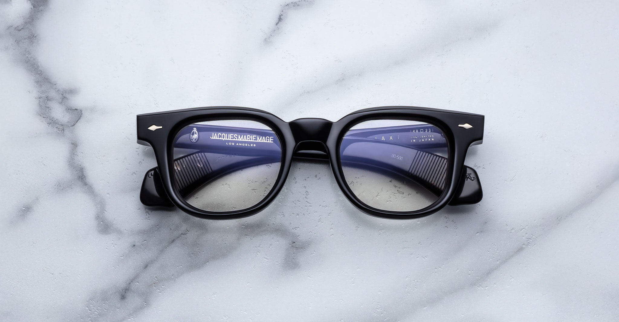 The Jacques Marie Mage Jax 2 black rectangular eyeglasses with clear lenses are displayed face up on a white marble surface, with the lenses reflecting nearby text and markings.