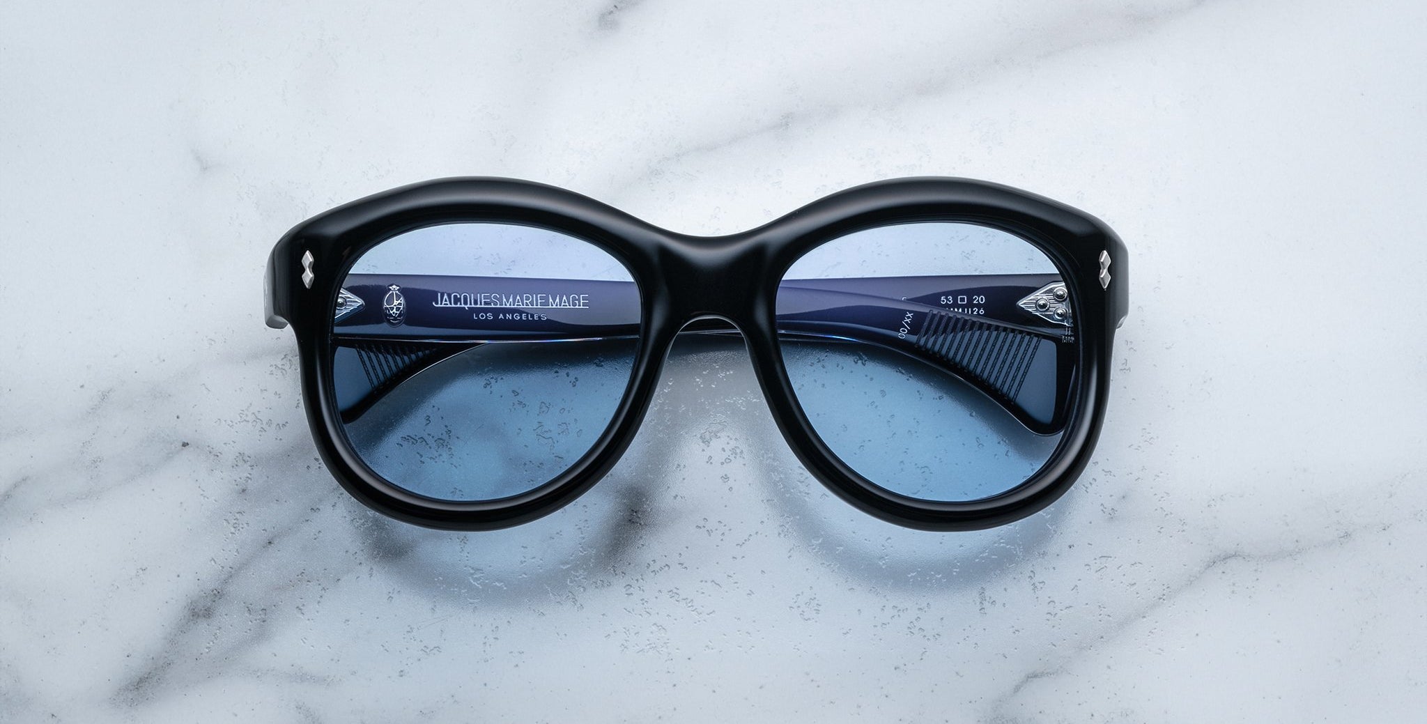 Jacques Marie Mage Jennie sunglasses with black frames and blue lenses, viewed from above.