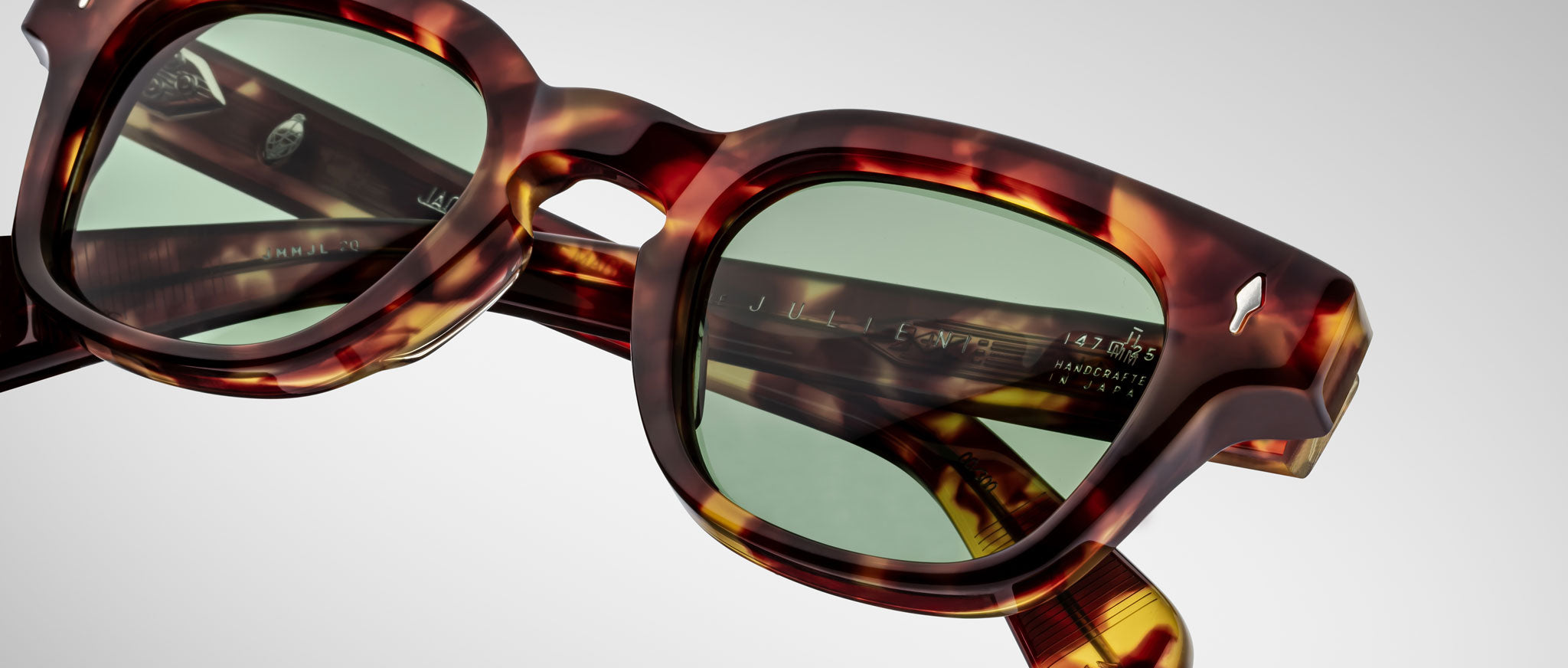 Close-up of Jacques Marie Mage Julien tortoiseshell sunglasses with green-tinted lenses, highlighting the frames detailed texture and visible inner-arm branding, set against a plain light background.