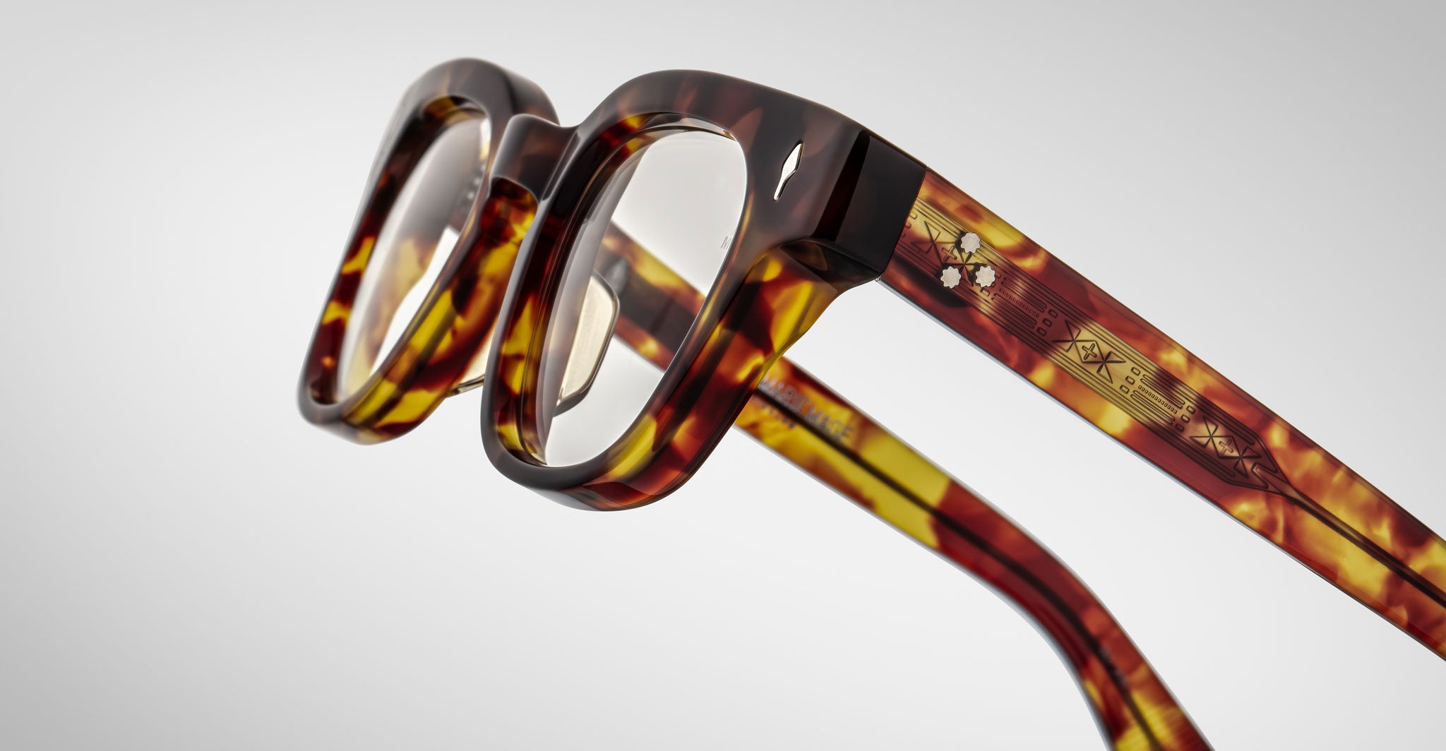 A close-up of Jacques Marie Mage Julien eyeglasses featuring thick square tortoiseshell frames, transparent lenses, and detailed multi-colored geometric patterns on the temples, set against a plain light background.