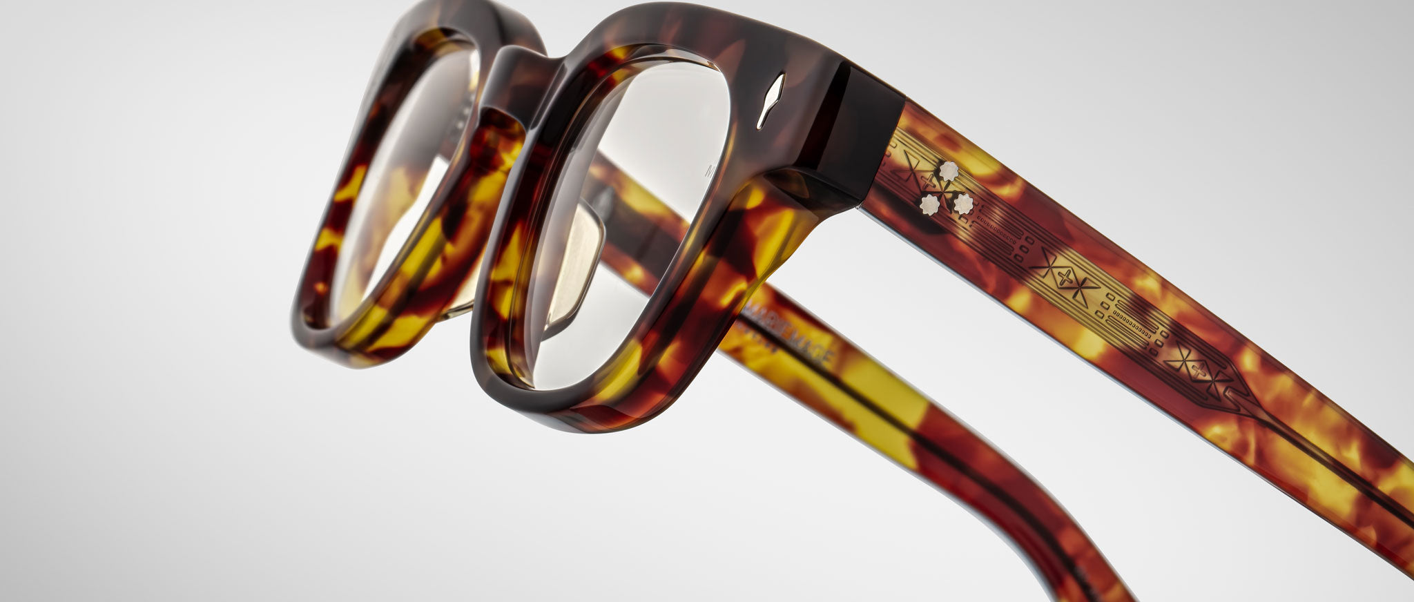 A close-up of Jacques Marie Mage Julien eyeglasses featuring thick square tortoiseshell frames, transparent lenses, and detailed multi-colored geometric patterns on the temples, set against a plain light background.