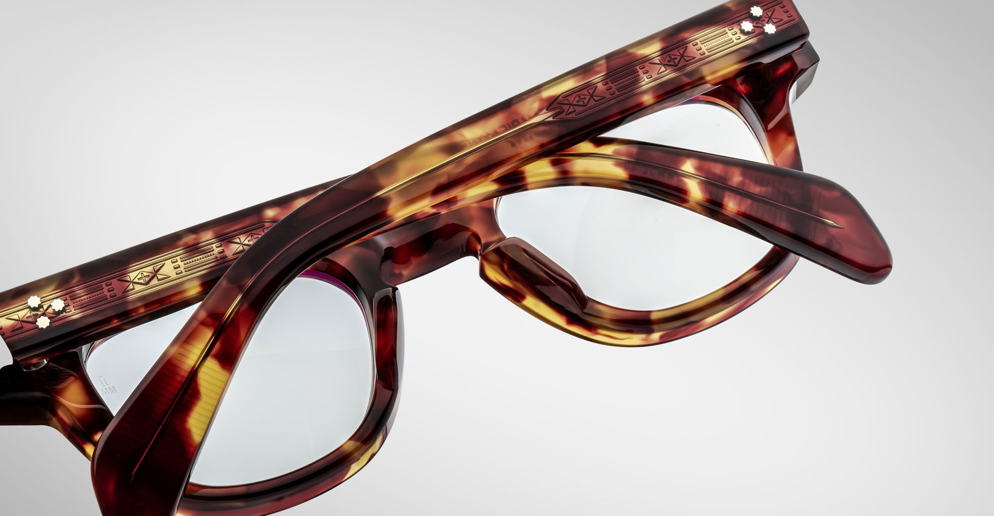 A close-up of two pairs of Jacques Marie Mage Julien tortoiseshell eyeglasses, one stacked atop the other, featuring gold and red accents on the arms against a plain, light background.