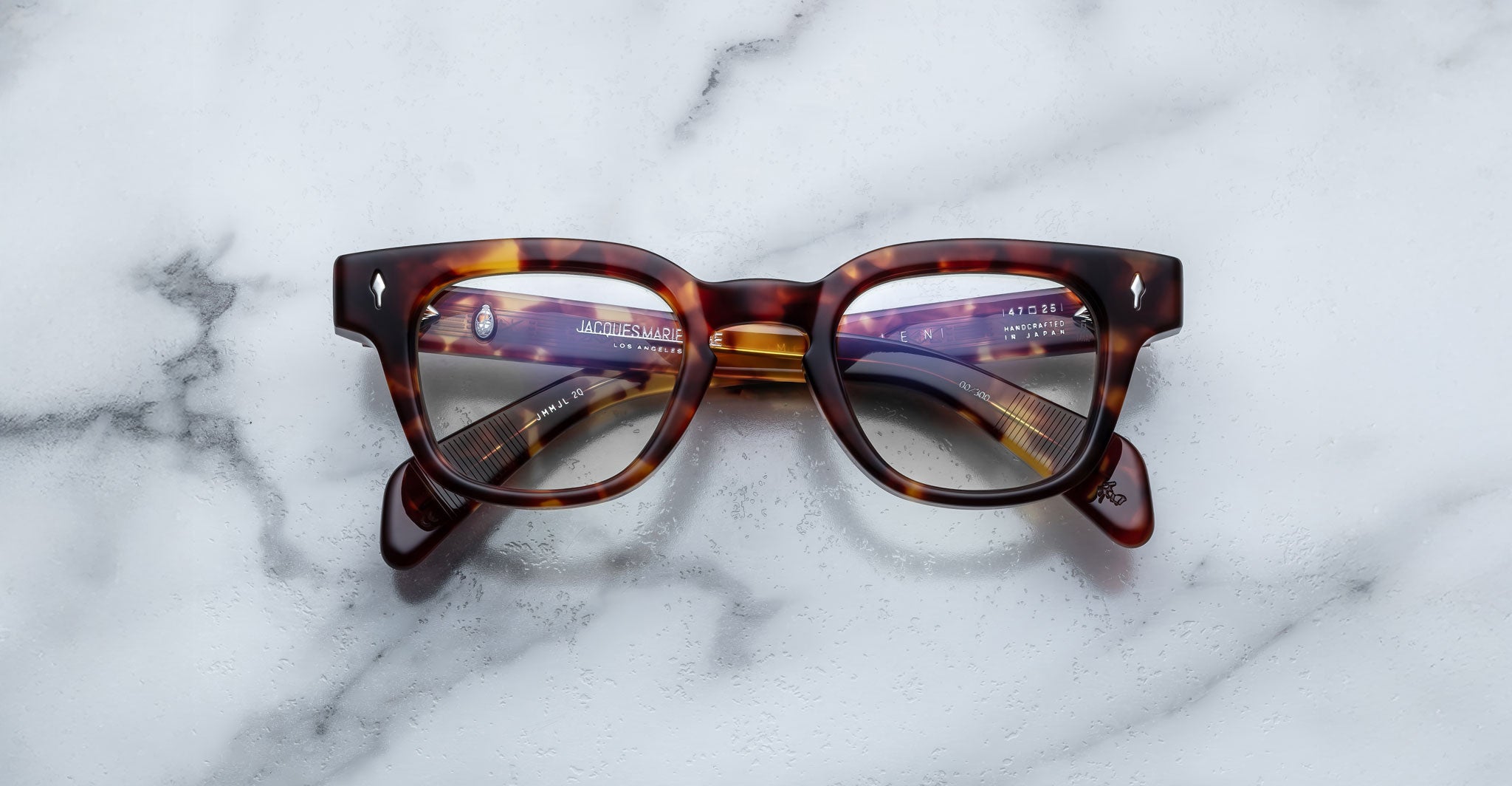 A pair of Jacques Marie Mage Julien tortoiseshell eyeglasses with thick frames rests on a white marble surface, their lenses reflecting text and objects.
