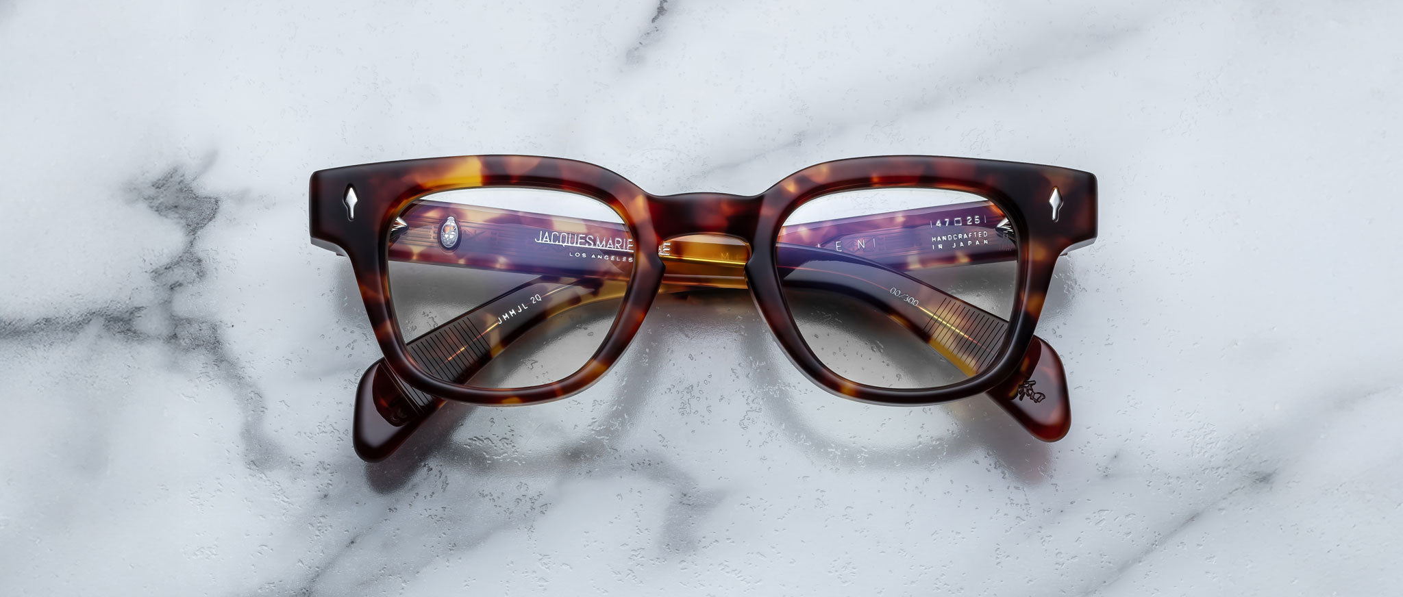 A pair of Jacques Marie Mage Julien tortoiseshell eyeglasses with thick frames rests on a white marble surface, their lenses reflecting text and objects.