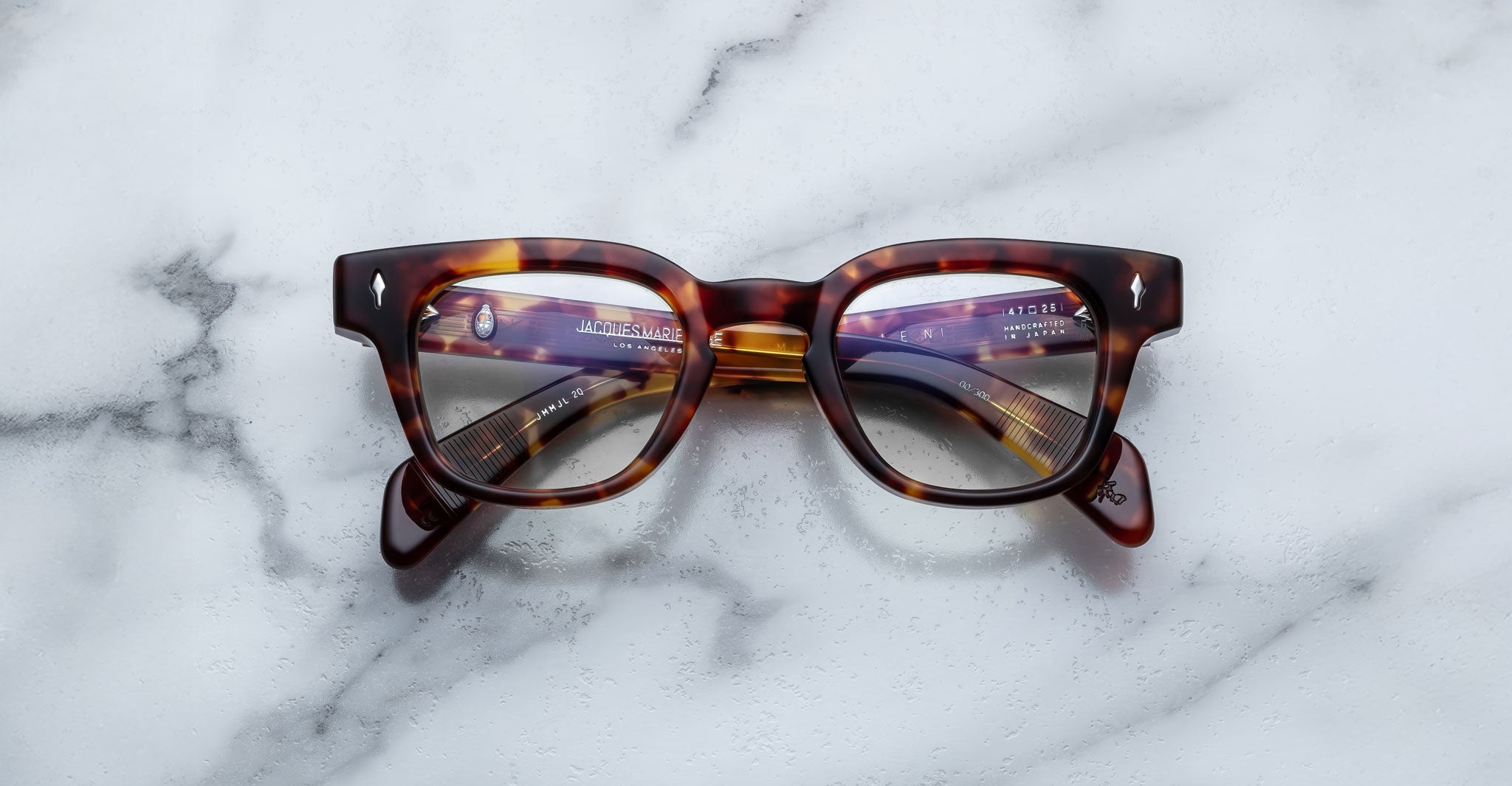 A pair of Jacques Marie Mage Julien tortoiseshell eyeglasses with thick frames rests on a white marble surface, their lenses reflecting text and objects.