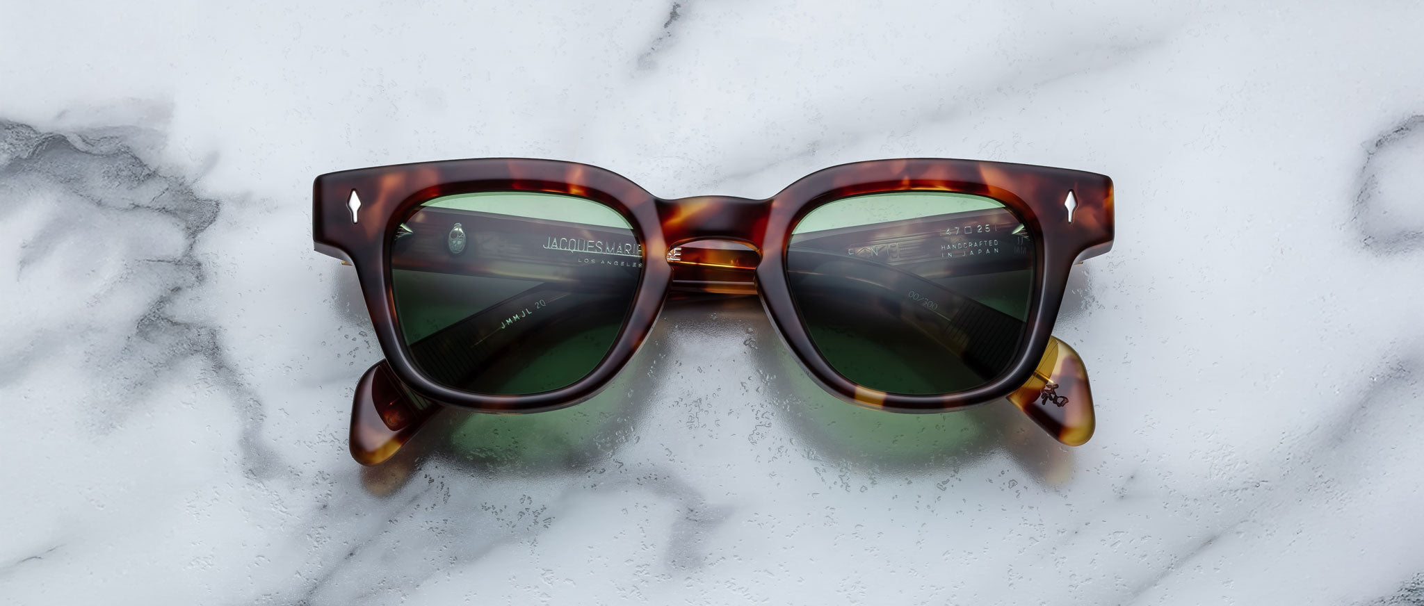 A pair of Jacques Marie Mage Julien tortoiseshell sunglasses with dark green lenses rests on a white marble surface.