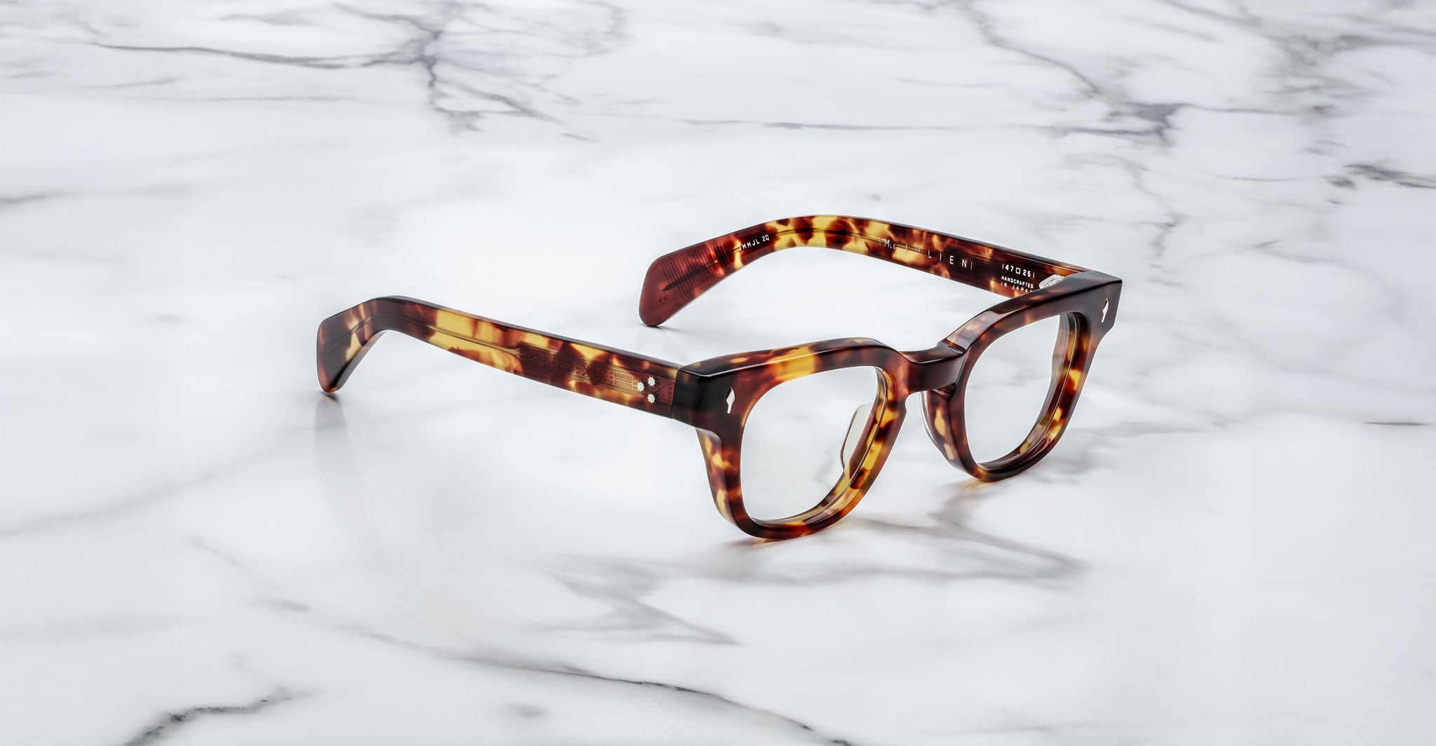 A pair of Jacques Marie Mage Julien eyeglasses with tortoiseshell thick frames rests on a white marble surface with gray veining.