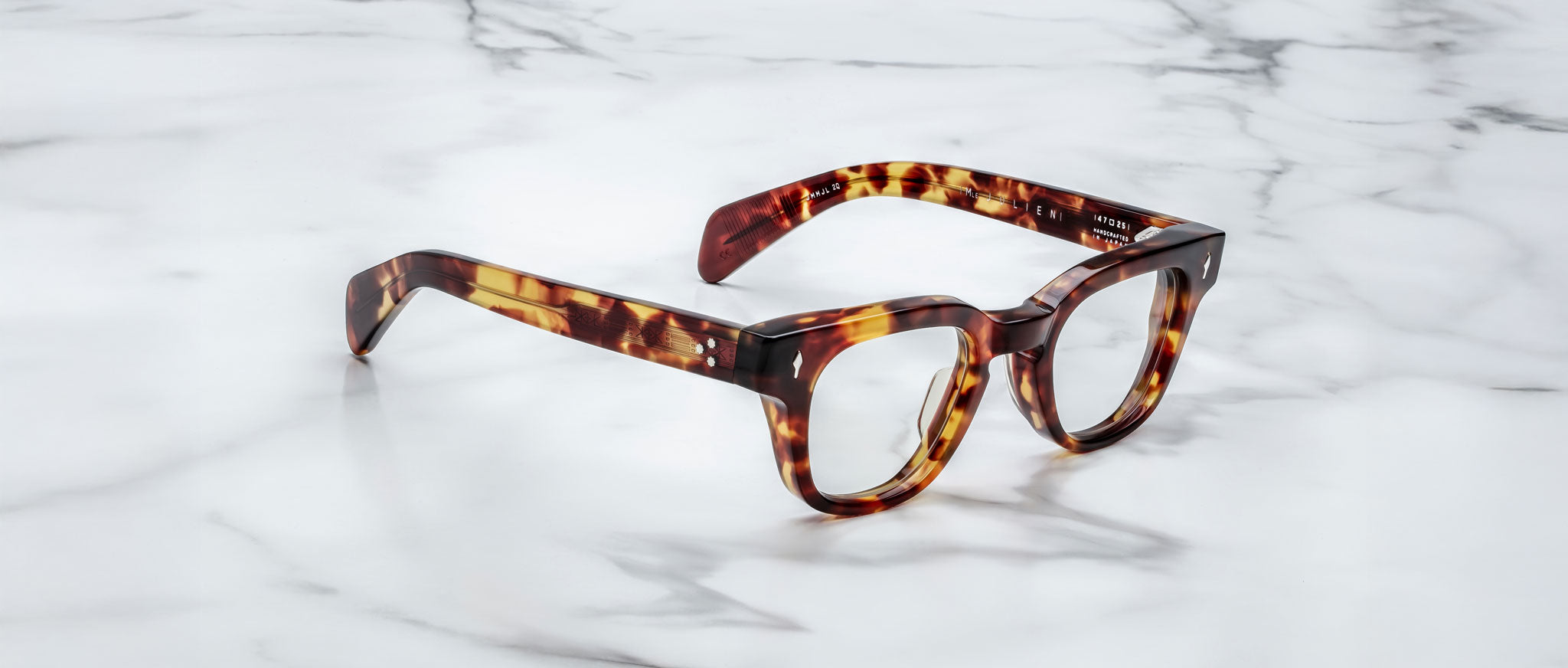A pair of Jacques Marie Mage Julien eyeglasses with tortoiseshell thick frames rests on a white marble surface with gray veining.