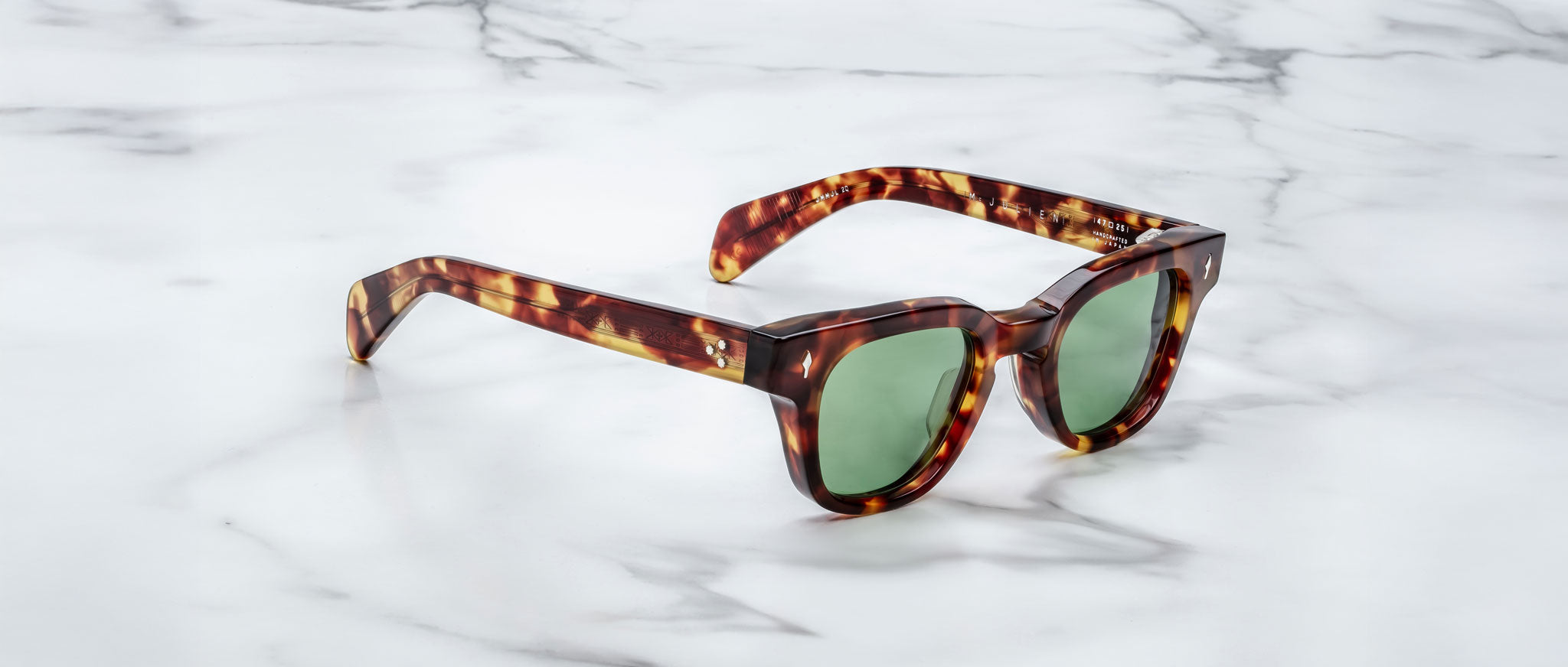 A pair of Jacques Marie Mage Julien tortoiseshell sunglasses with green lenses rests on a white marble surface with gray veining.