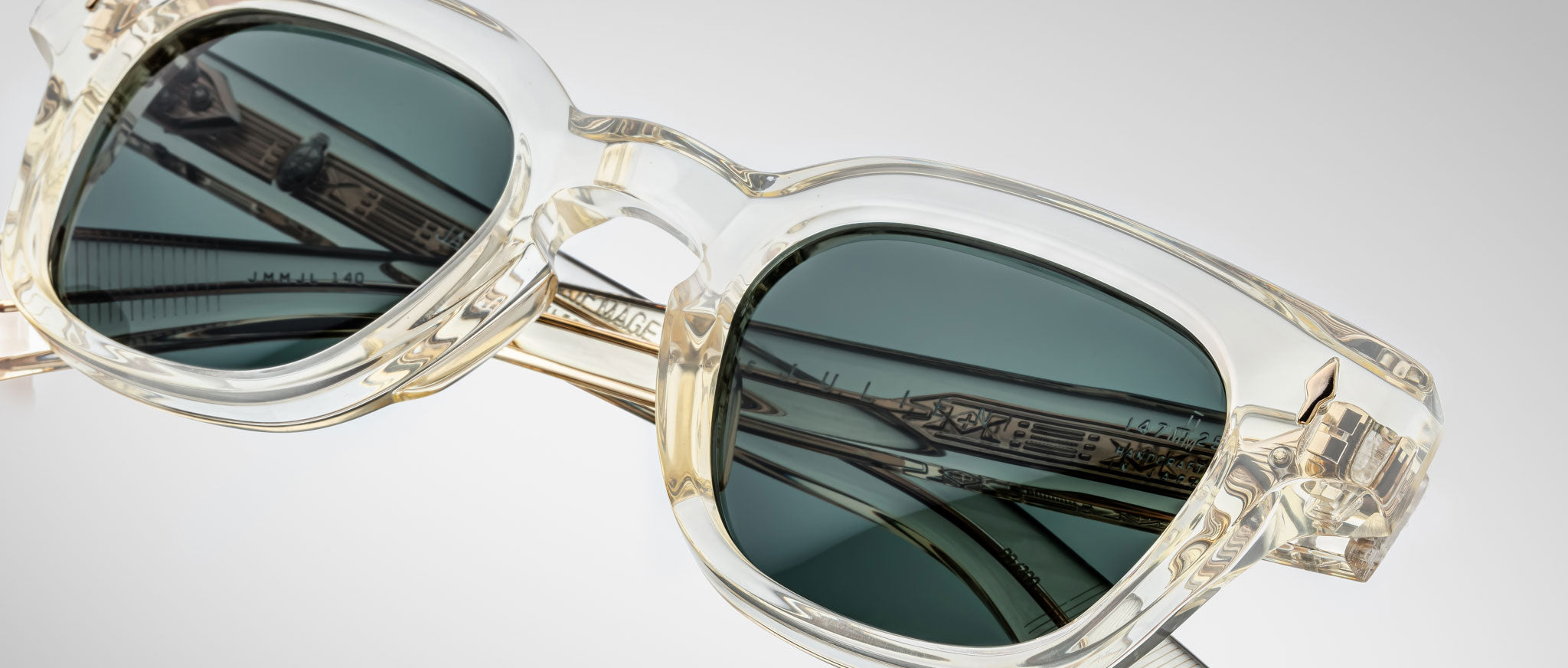 Close-up of Jacques Marie Mage Julien sunglasses with translucent rectangular frames, dark lenses, and arms featuring metallic detailing and engraved text, all set against a plain light background.