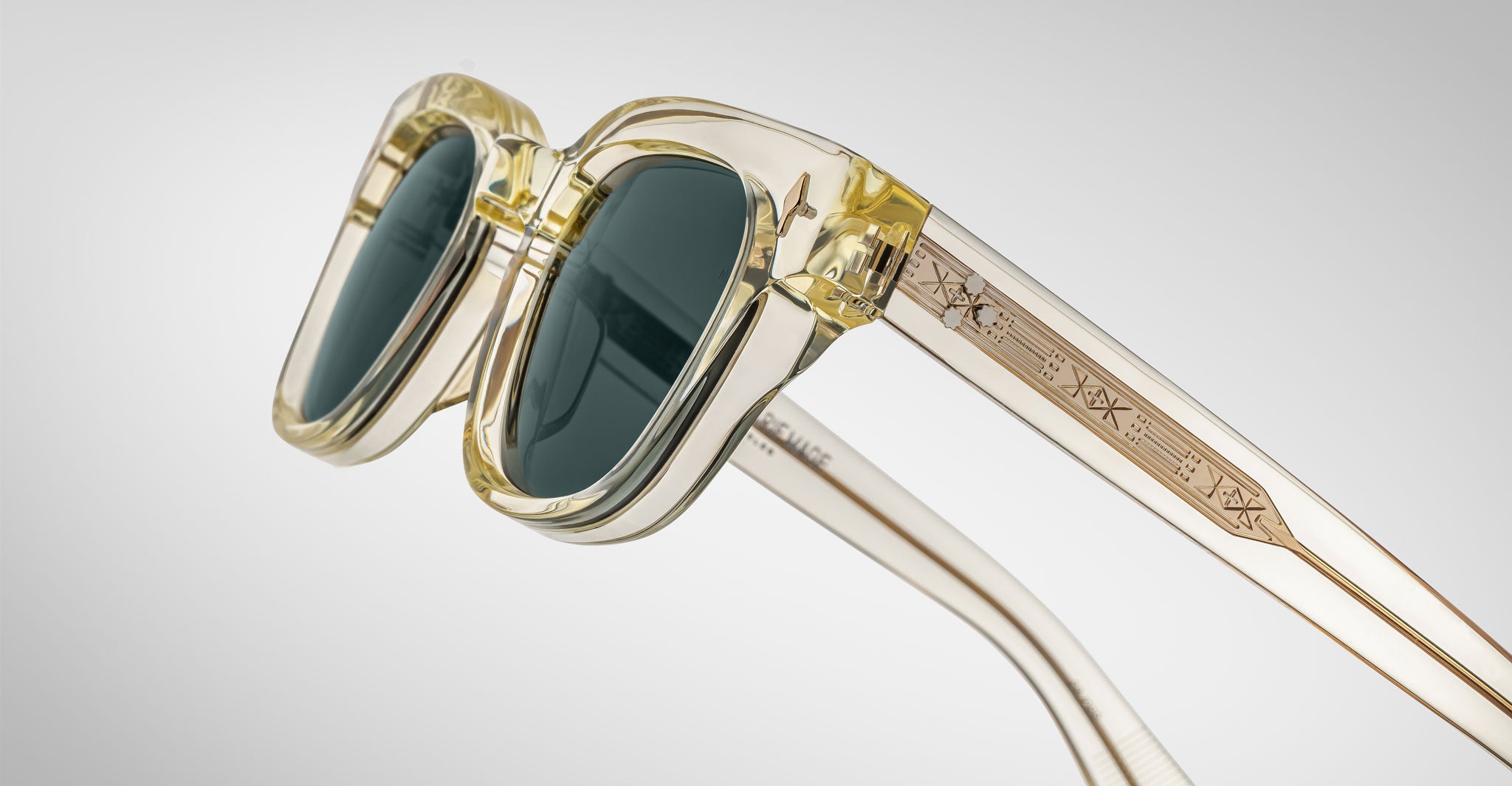 A close-up of Jacques Marie Mage Julien translucent yellow sunglasses with dark lenses and geometric engravings on the arms, set against a plain light gray background.