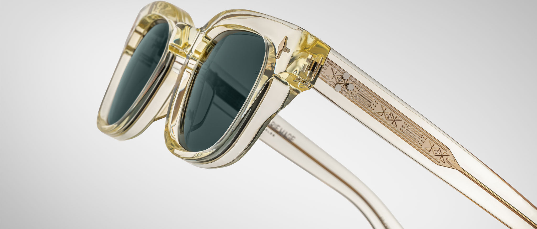 A close-up of Jacques Marie Mage Julien translucent yellow sunglasses with dark lenses and geometric engravings on the arms, set against a plain light gray background.