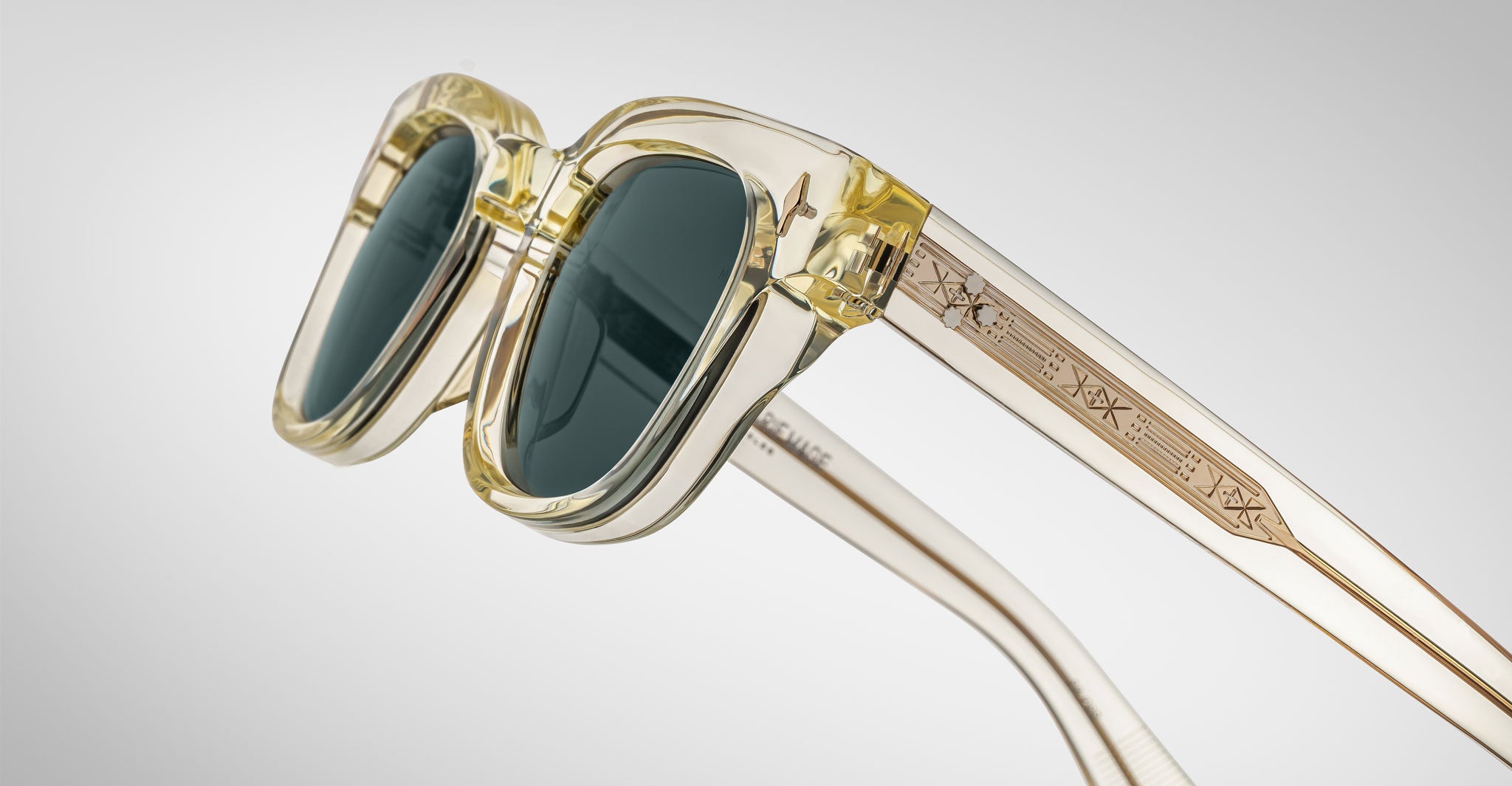 A close-up of Jacques Marie Mage Julien translucent yellow sunglasses with dark lenses and geometric engravings on the arms, set against a plain light gray background.