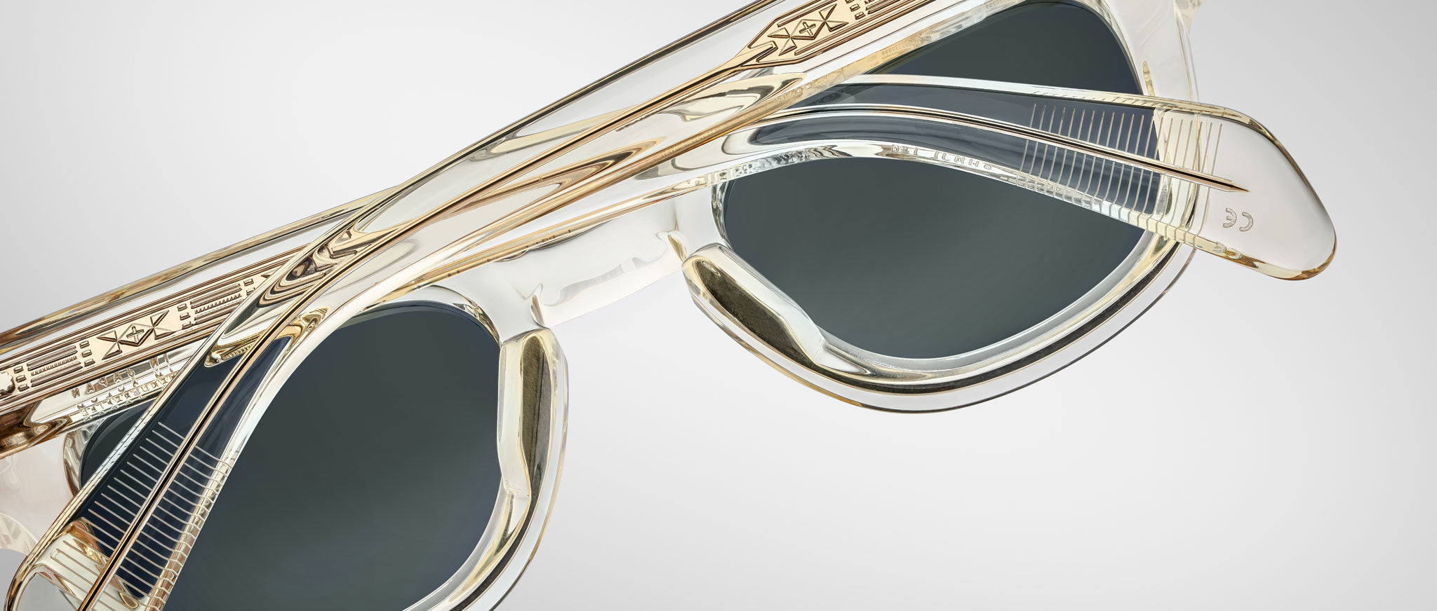 Close-up of Jacques Marie Mage Julien transparent sunglasses with gold accents and dark lenses, highlighting the intricate patterns on the frames and temples against a light gray background.