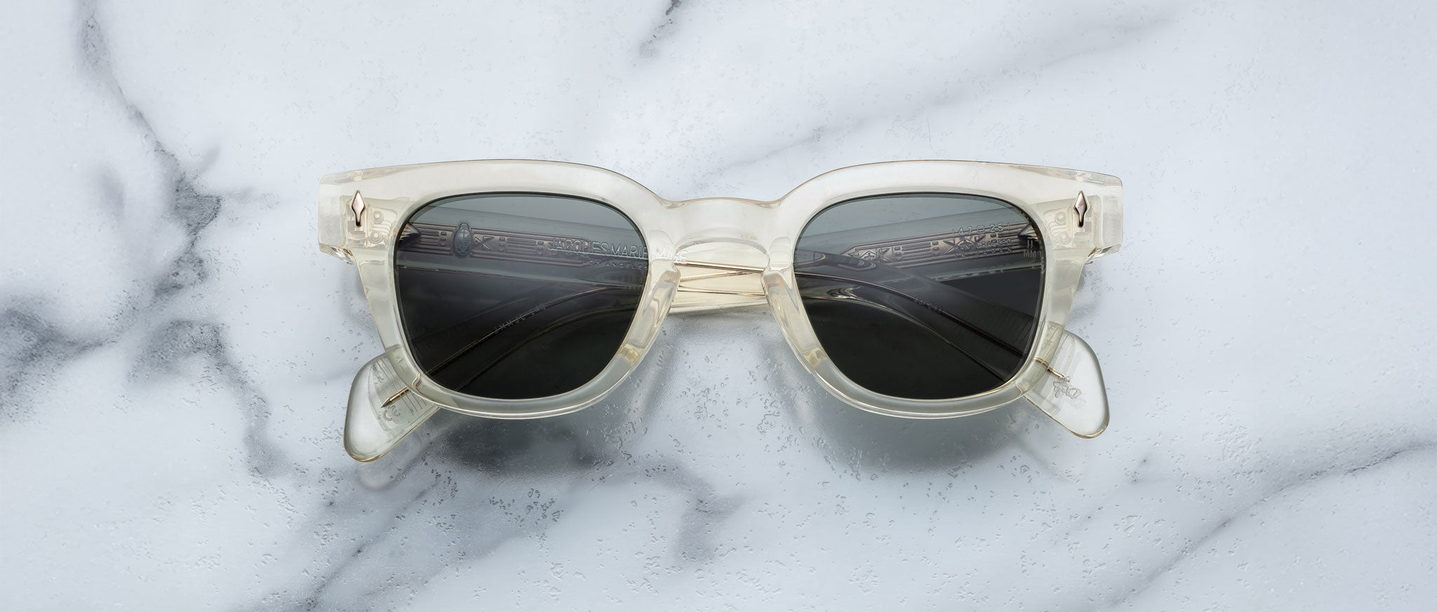 The Jacques Marie Mage Julien sunglasses, featuring clear frames and dark lenses, are shown face up with folded arms on a white marble surface with gray veining.