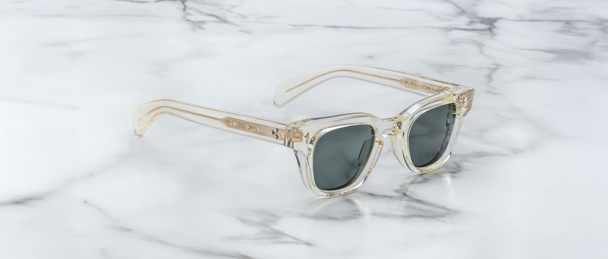 A pair of Jacques Marie Mage Julien sunglasses with clear frames and dark lenses rests on white marble with gray veining. The temples and frame edges feature a subtle beige or gold tint.