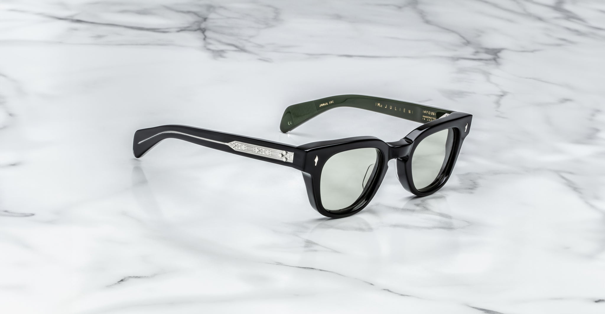 The Jacques Marie Mage Julien sunglasses, featuring thick black frames, green-tinted lenses, and silver-detailed temples with inner branding, rest on a white marble surface.