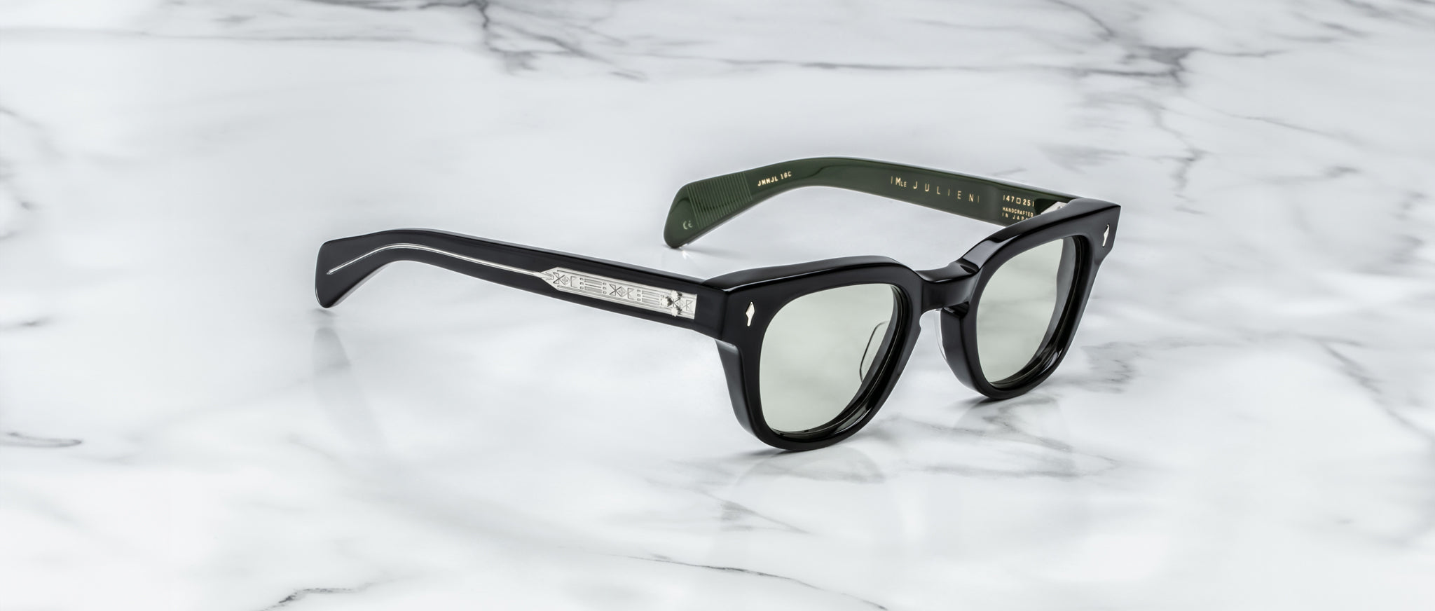 The Jacques Marie Mage Julien sunglasses, featuring thick black frames, green-tinted lenses, and silver-detailed temples with inner branding, rest on a white marble surface.