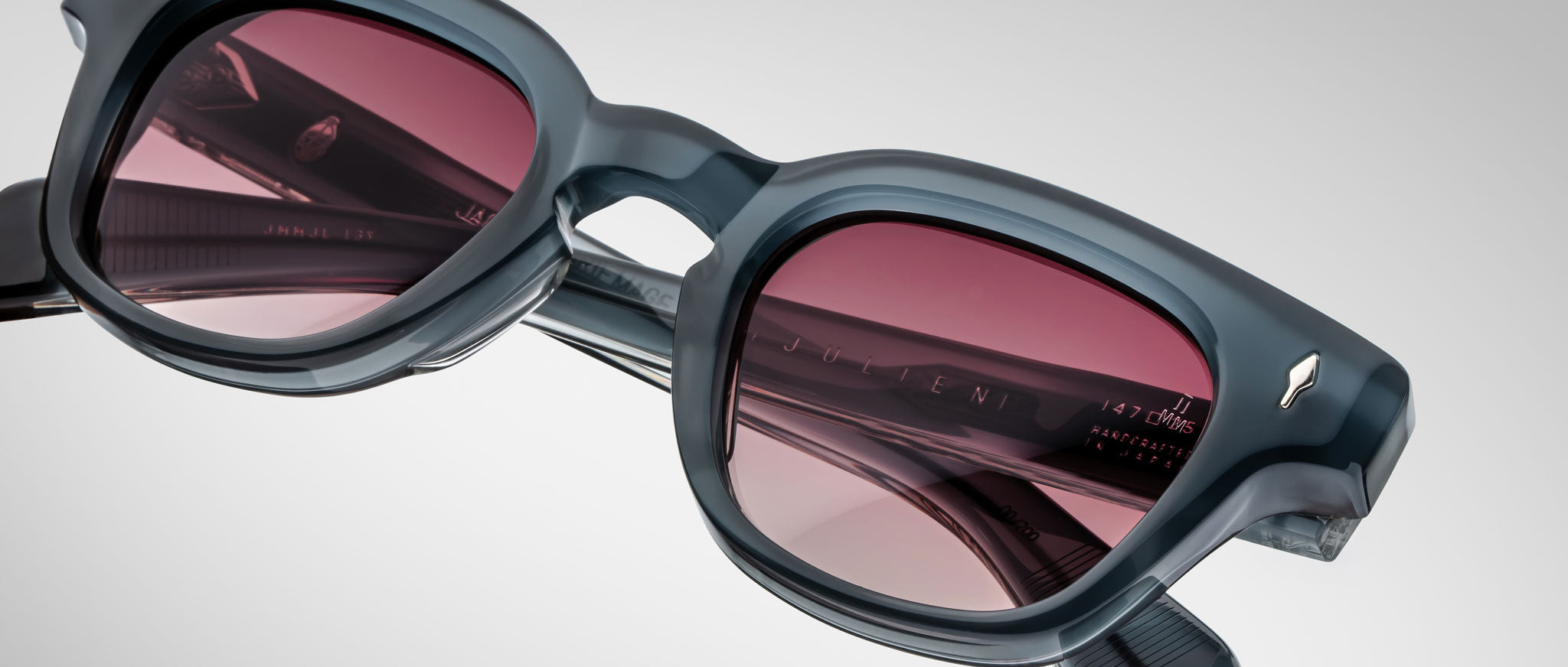 A close-up of Jacques Marie Mage Julien sunglasses in dark gray acetate with thick frames and red-tinted lenses, highlighting the hinge detail and reflective logo on the temple arm against a light background.