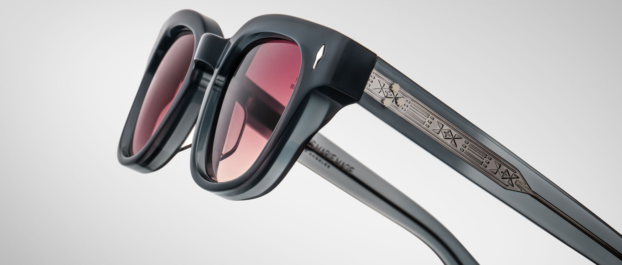 A close-up of Jacques Marie Mage Julien sunglasses featuring thick black frames, tinted red lenses, and an intricate metallic pattern on the temples, set against a light gray background.