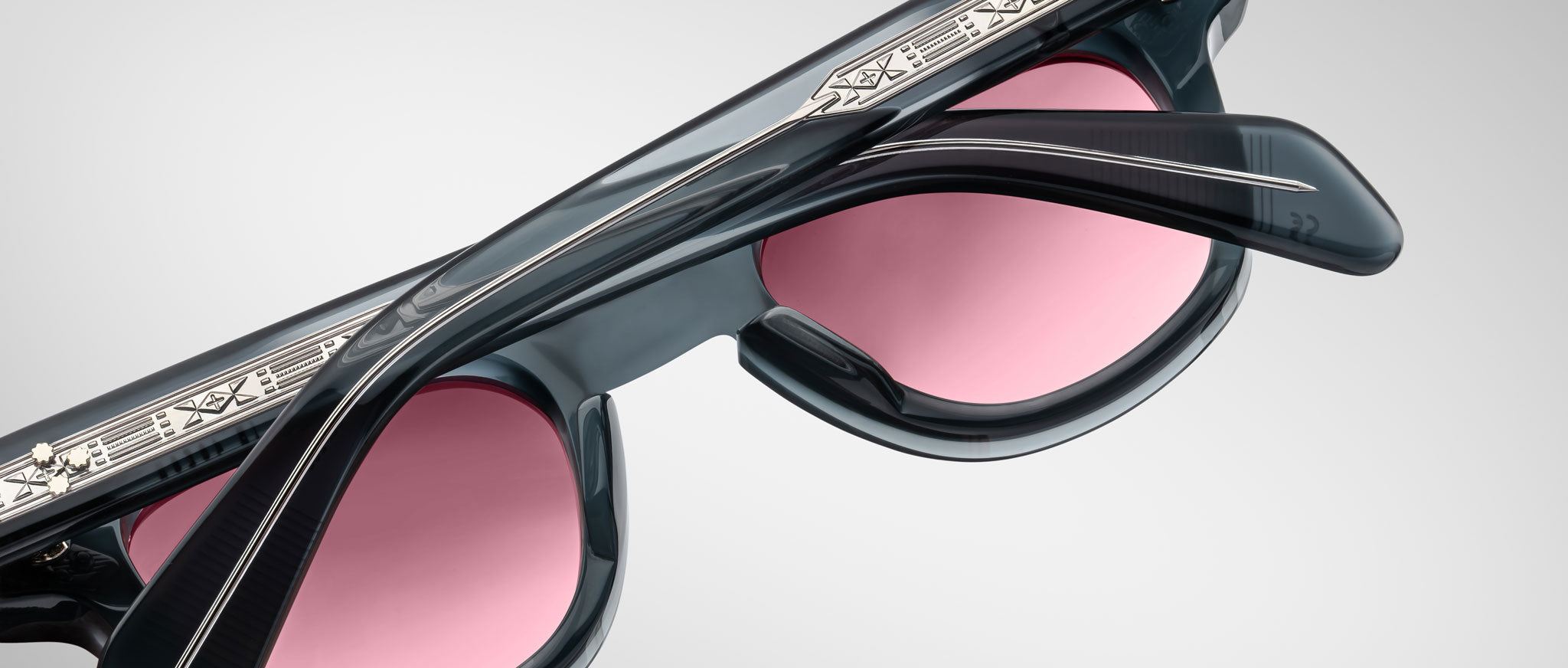 Close-up of Jacques Marie Mage Julien sunglasses in black with pink-tinted lenses, featuring intricate silver engravings on the top frame and textured temples, set against a plain light gray background.