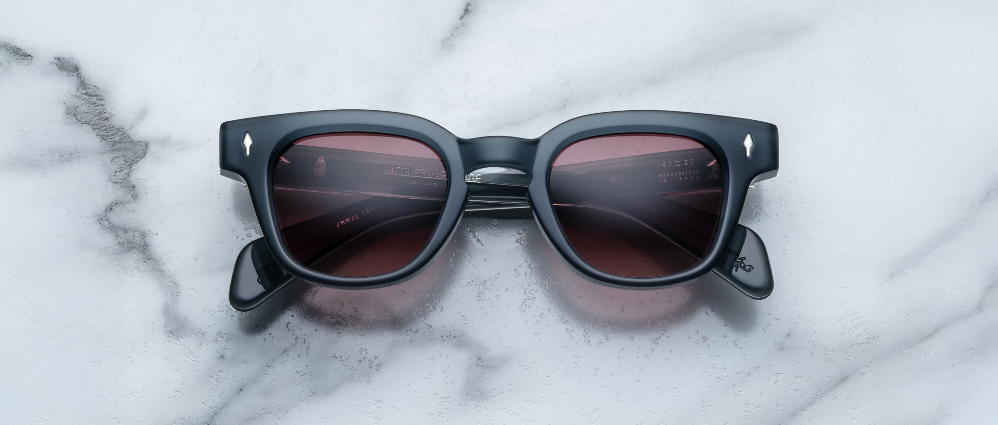 The Jacques Marie Mage Julien sunglasses, featuring black frames and dark reddish-brown lenses, rest on a white marble surface with grey veining. Viewed from above, the arms of the glasses are partially visible.