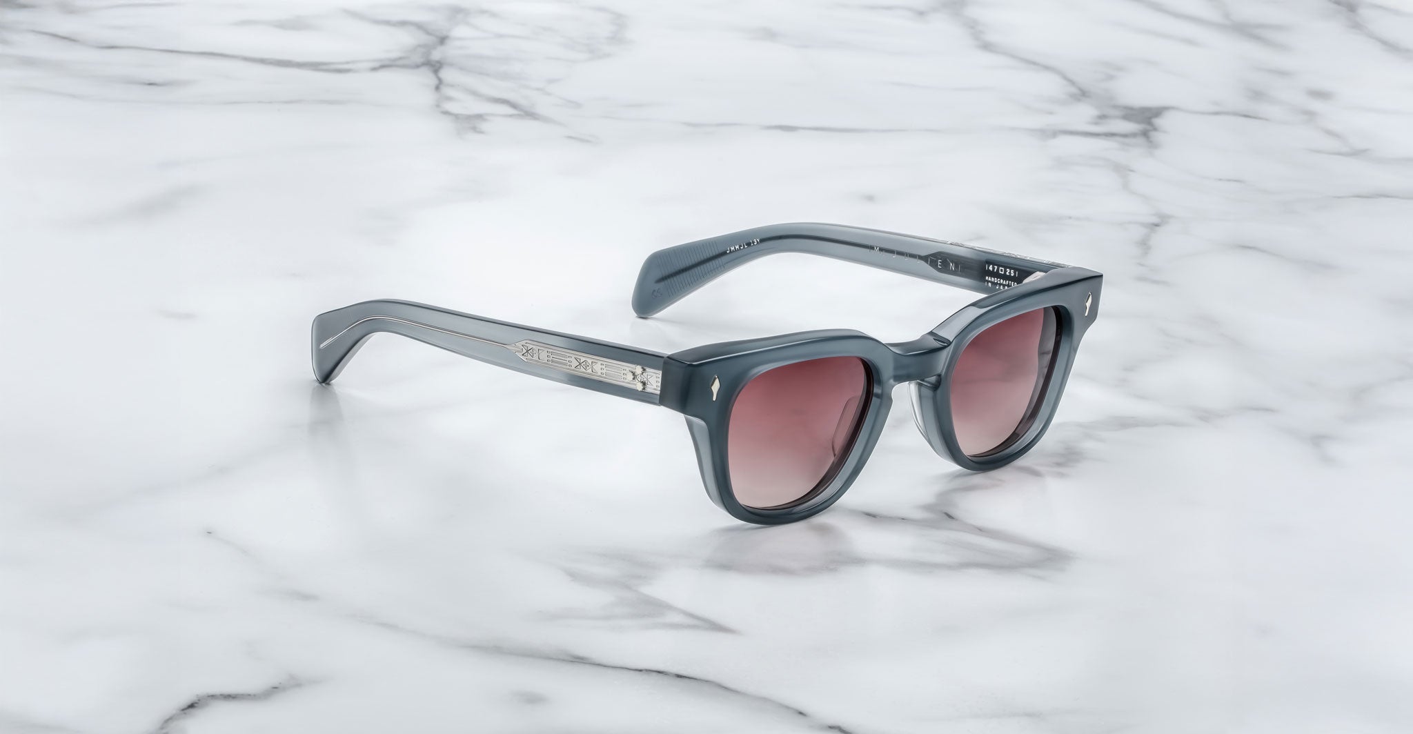 The Jacques Marie Mage Julien sunglasses feature a dark gray rectangular frame, tinted lenses, and silver accents on the arms, displayed on a white marble surface.