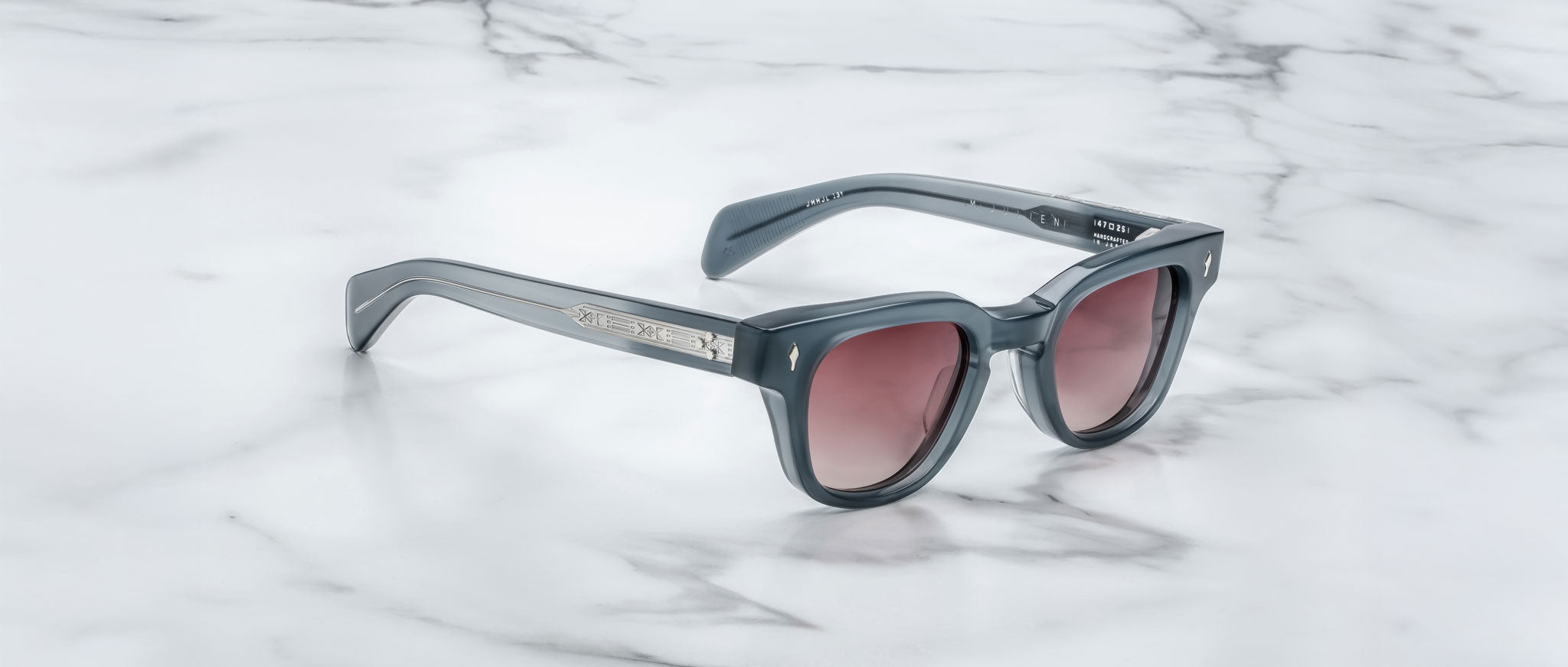 The Jacques Marie Mage Julien sunglasses feature a dark gray rectangular frame, tinted lenses, and silver accents on the arms, displayed on a white marble surface.