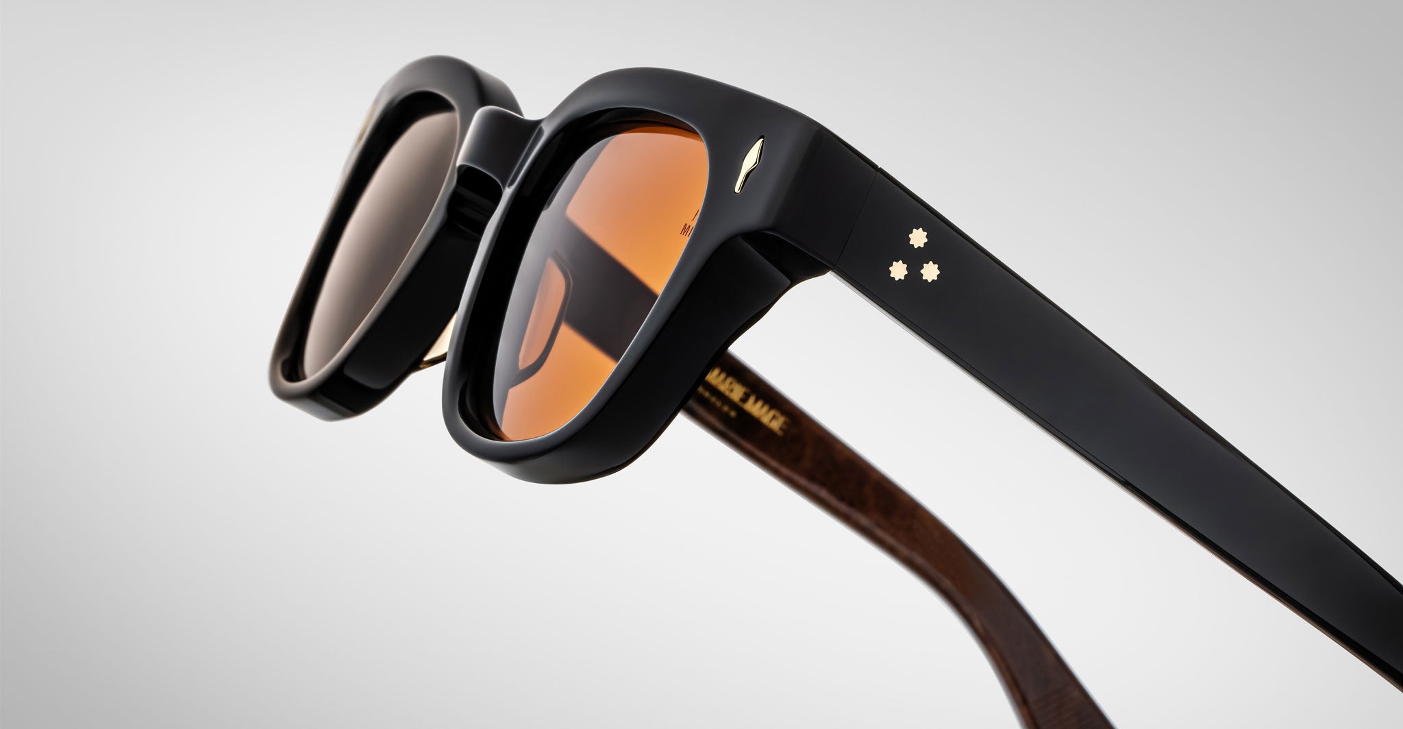 Close-up of Jacques Marie Mage Julien sunglasses with thick black frames, orange-tinted lenses, and gold star details on the temples. The inner arms feature a wood-like texture and gold lettering.