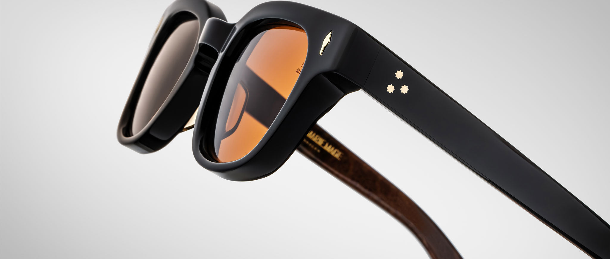 Close-up of Jacques Marie Mage Julien sunglasses with thick black frames, orange-tinted lenses, and gold star details on the temples. The inner arms feature a wood-like texture and gold lettering.