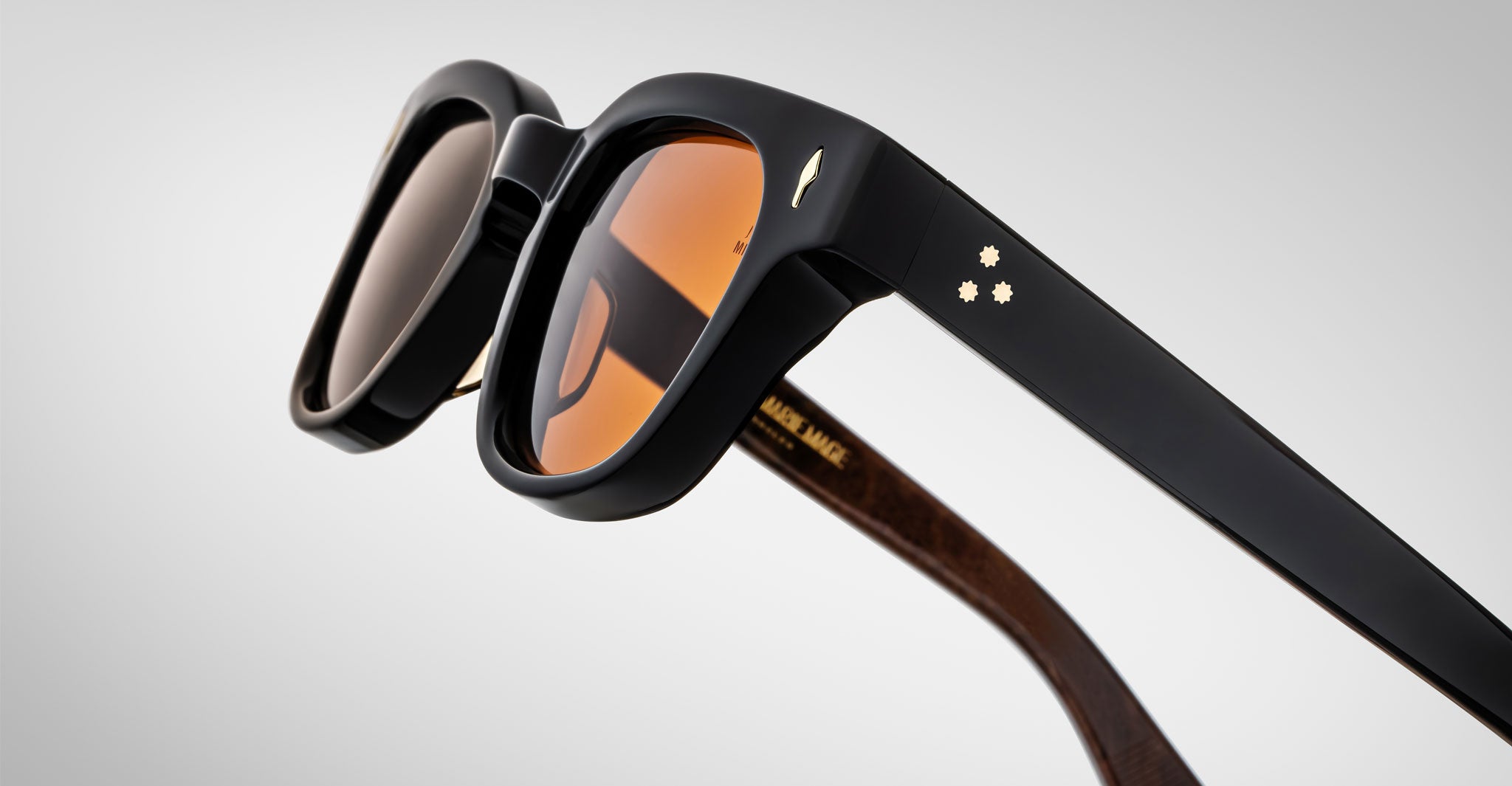 Close-up of Jacques Marie Mage Julien sunglasses with thick black frames, orange-tinted lenses, and gold star details on the temples. The inner arms feature a wood-like texture and gold lettering.