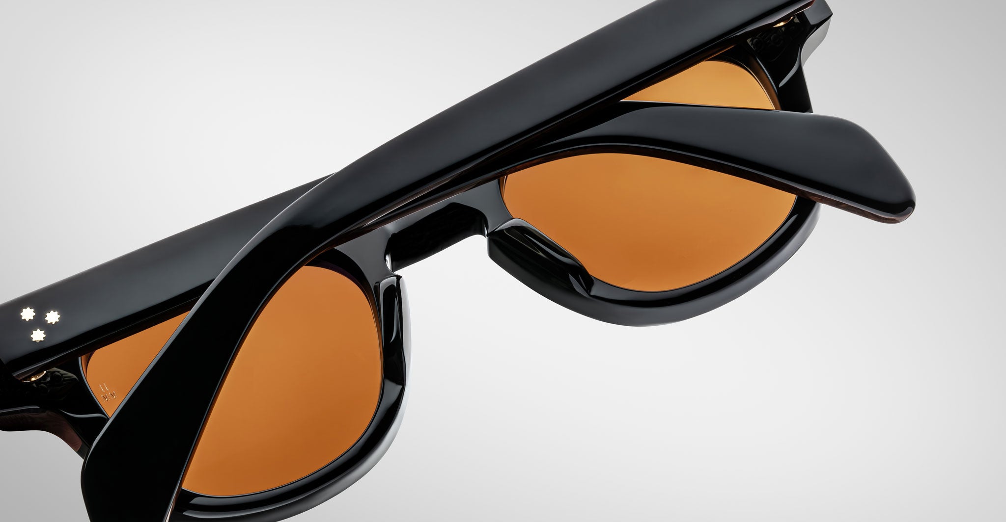 Close-up of Jacques Marie Mage Julien black sunglasses with amber lenses, partially folded, against a plain light gray background. Three small white dots appear on one arm.