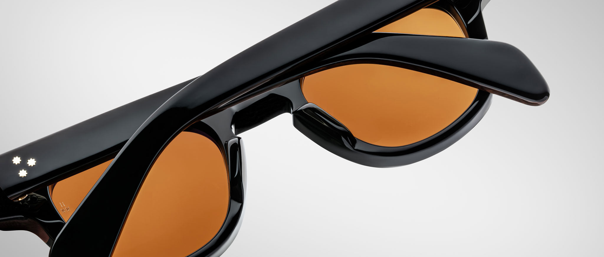 Close-up of Jacques Marie Mage Julien black sunglasses with amber lenses, partially folded, against a plain light gray background. Three small white dots appear on one arm.