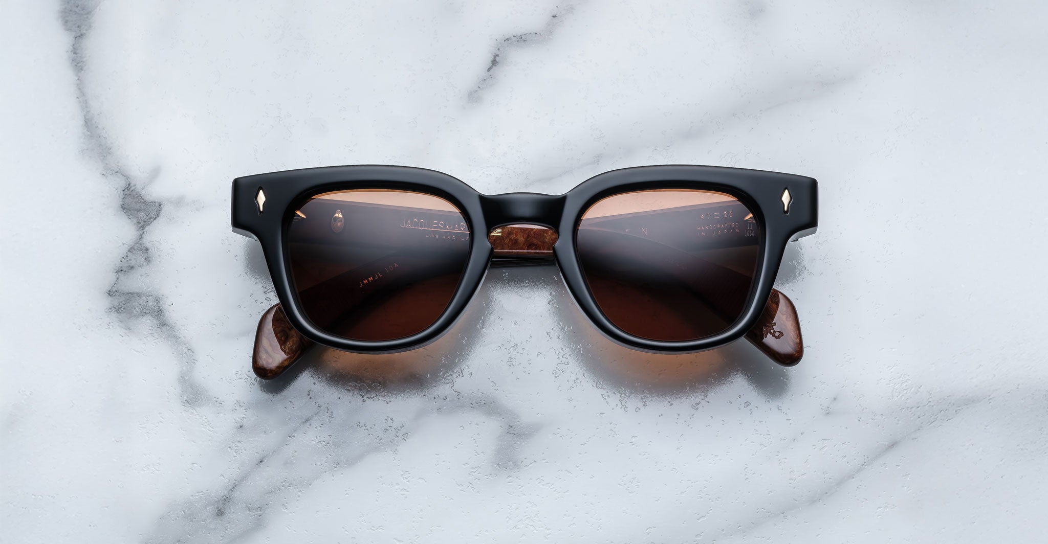 The Jacques Marie Mage Julien features black rectangular frames, brown-tinted lenses, and marbled brown arms, elegantly displayed on a white marble surface.