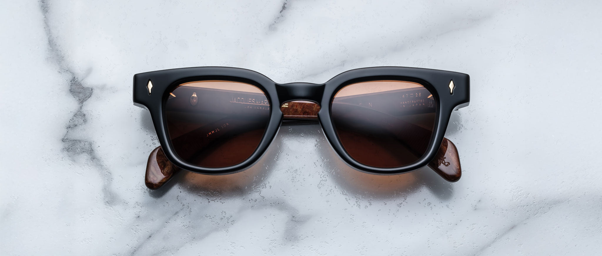 The Jacques Marie Mage Julien features black rectangular frames, brown-tinted lenses, and marbled brown arms, elegantly displayed on a white marble surface.
