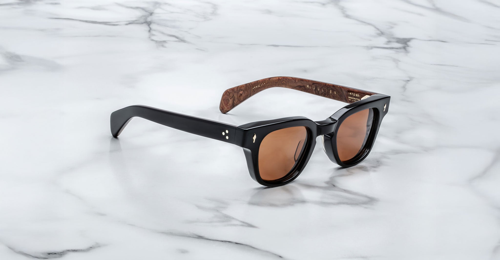 A pair of Jacques Marie Mage Julien black sunglasses with brown-tinted lenses and patterned brown inner arms rests on a white marble surface.