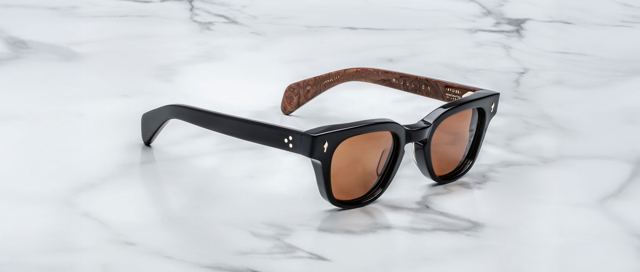 A pair of Jacques Marie Mage Julien black sunglasses with brown-tinted lenses and patterned brown inner arms rests on a white marble surface.