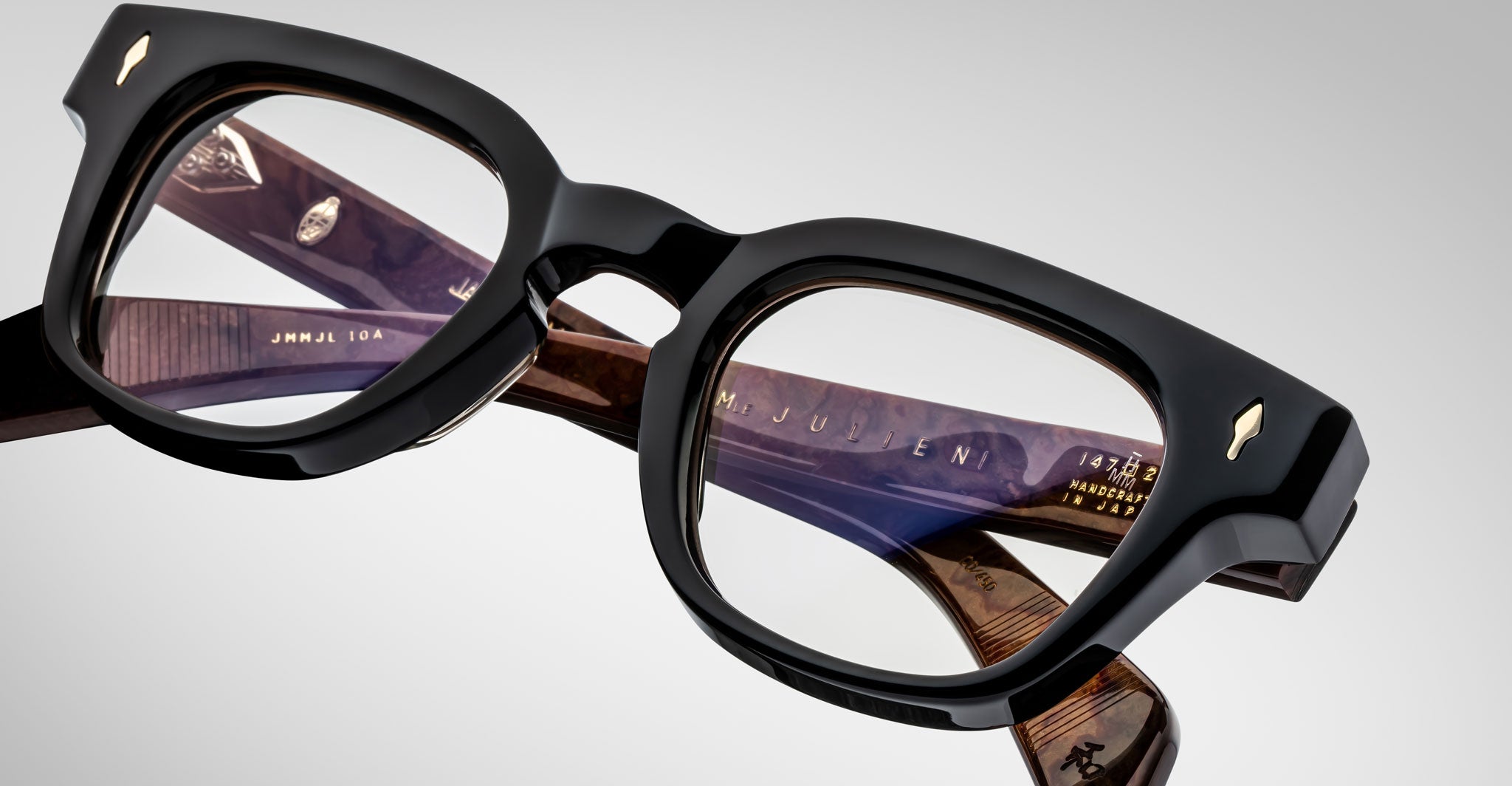 A close-up of Jacques Marie Mage Julien eyeglasses in black and brown with thick frames, clear lenses, and detailed temple markings, set against a plain gray background.