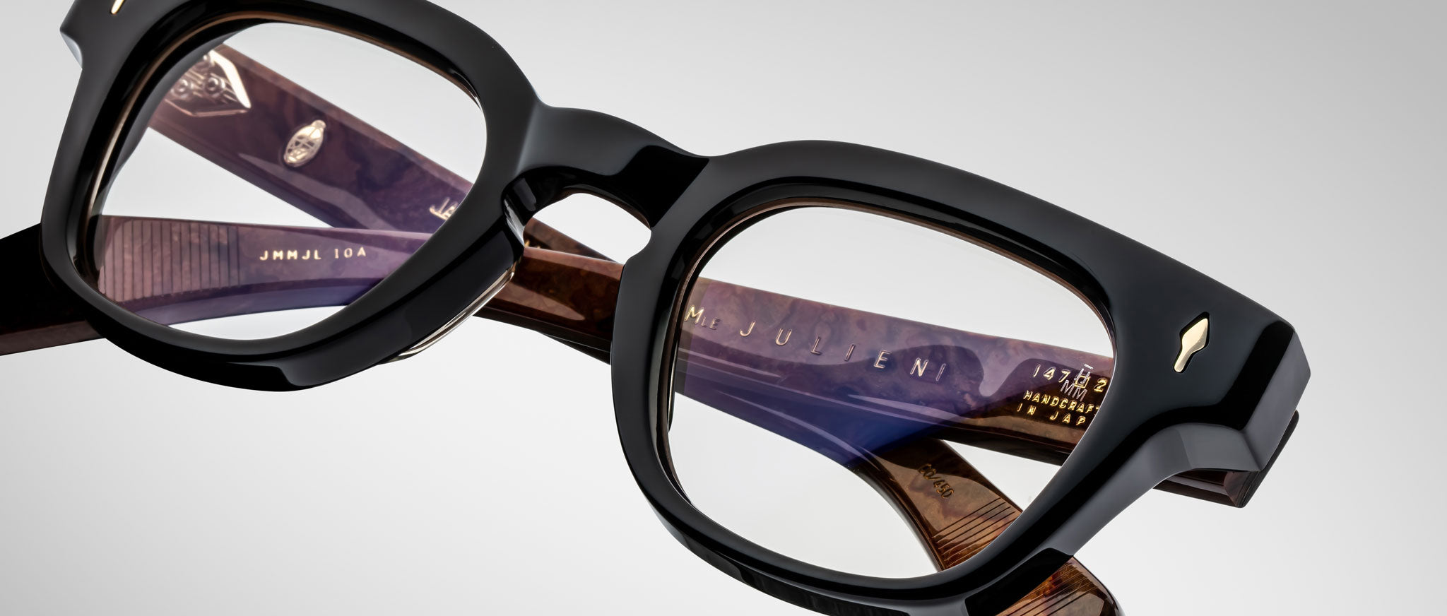 A close-up of Jacques Marie Mage Julien eyeglasses in black and brown with thick frames, clear lenses, and detailed temple markings, set against a plain gray background.