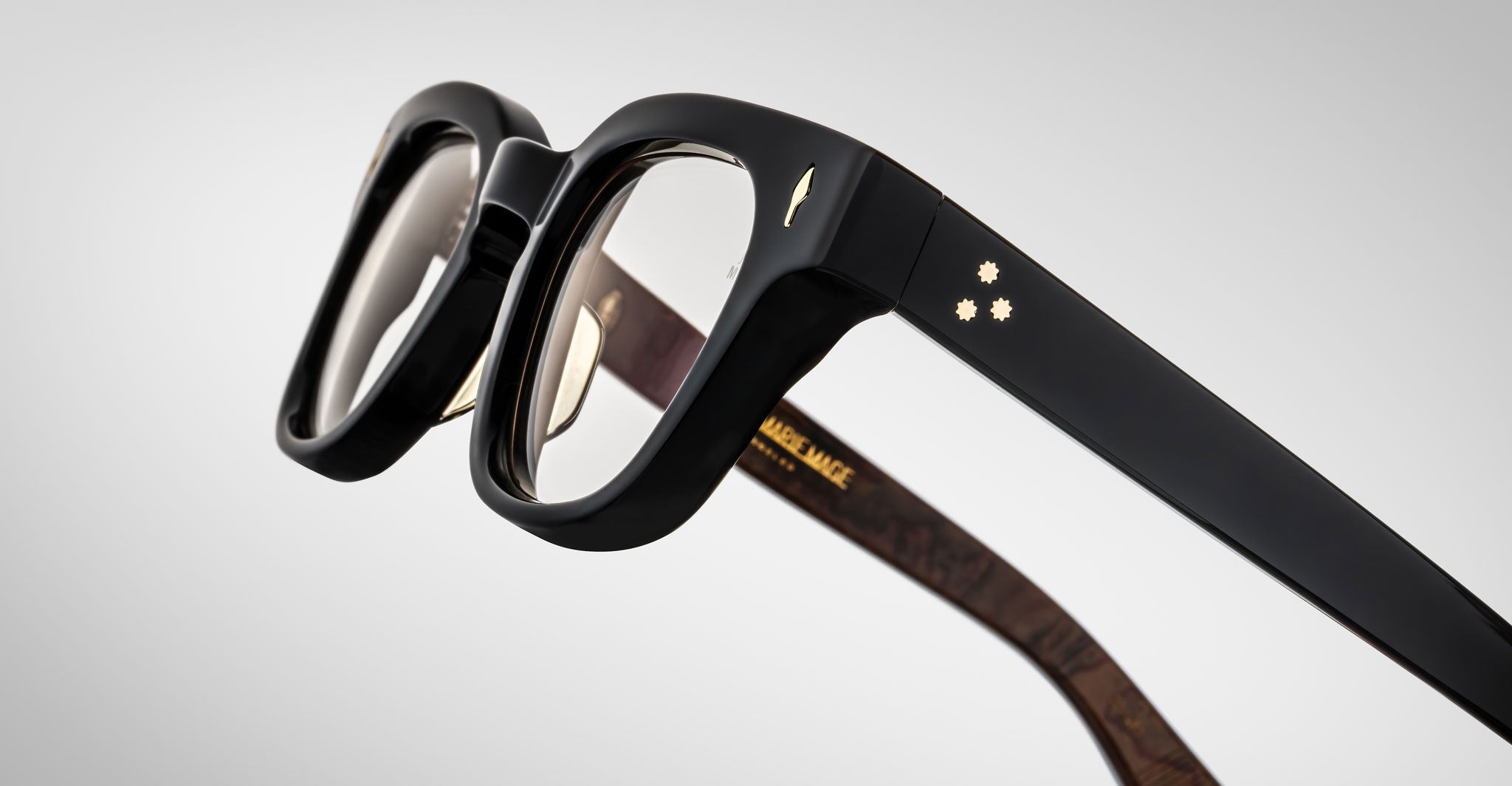 Close-up of Jacques Marie Mage Julien black eyeglass frames with thick rims. The temple has two small gold star details and a gold bird insignia, with gold text inside the arm. Background is light gray.