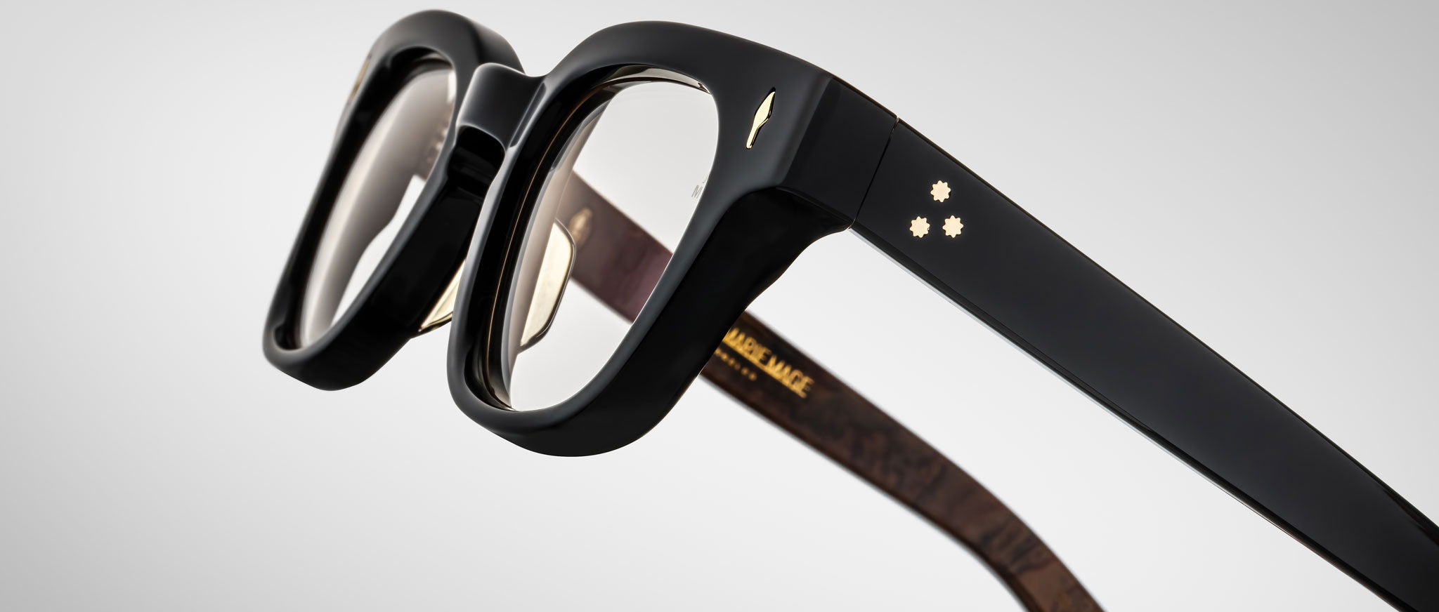 Close-up of Jacques Marie Mage Julien black eyeglass frames with thick rims. The temple has two small gold star details and a gold bird insignia, with gold text inside the arm. Background is light gray.