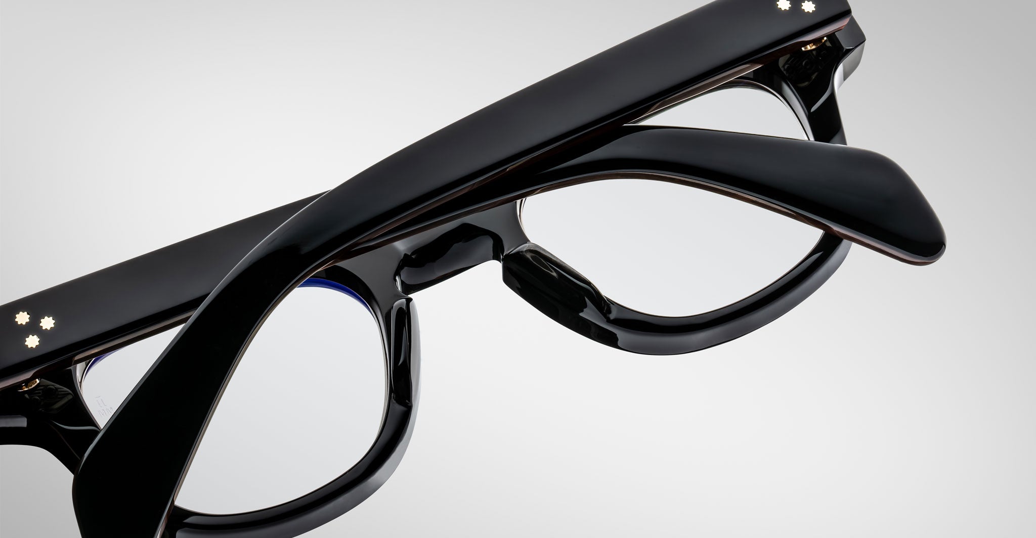 Close-up of Jacques Marie Mage Julien black eyeglasses with thick frames and temples, arms crossed over each other, set against a plain light gray background.