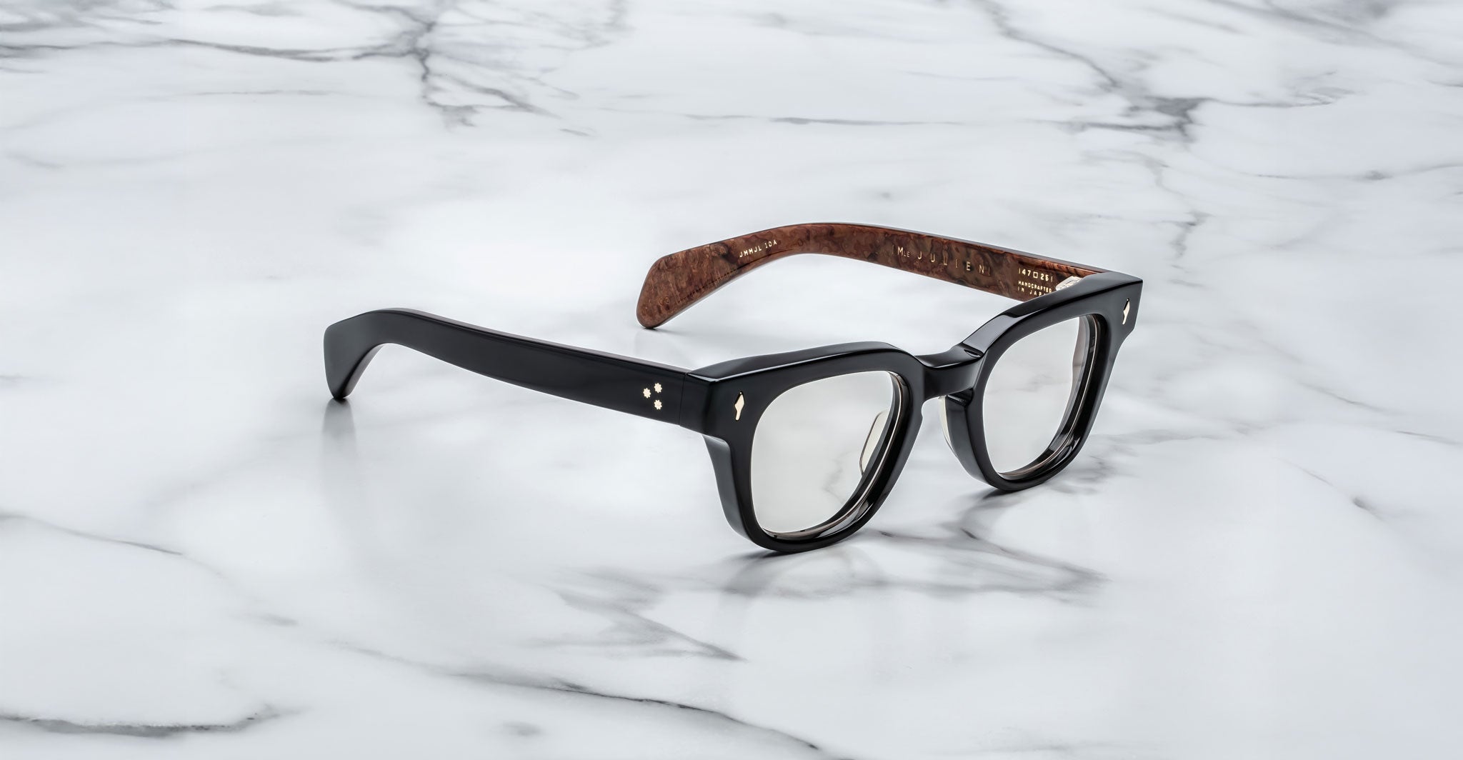 The Jacques Marie Mage Julien eyeglasses feature black rectangular glossy frames with brown inner arms, displayed on a white marble surface with gray veining.