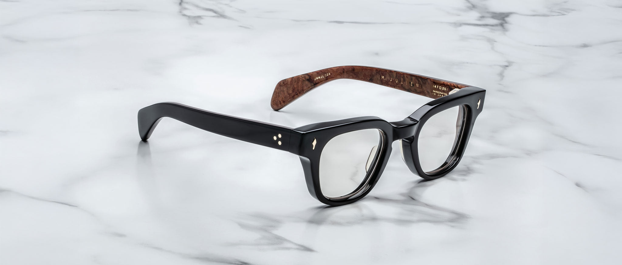 The Jacques Marie Mage Julien eyeglasses feature black rectangular glossy frames with brown inner arms, displayed on a white marble surface with gray veining.