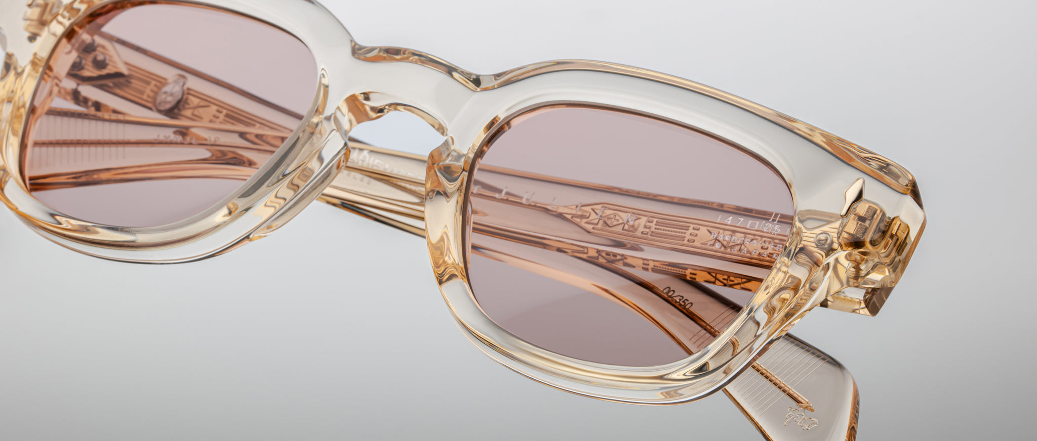 Close-up of Jacques Marie Mage Julien transparent beige rectangular eyeglasses with light brown tinted lenses and detailed engravings on the temples, set against a light gray background.
