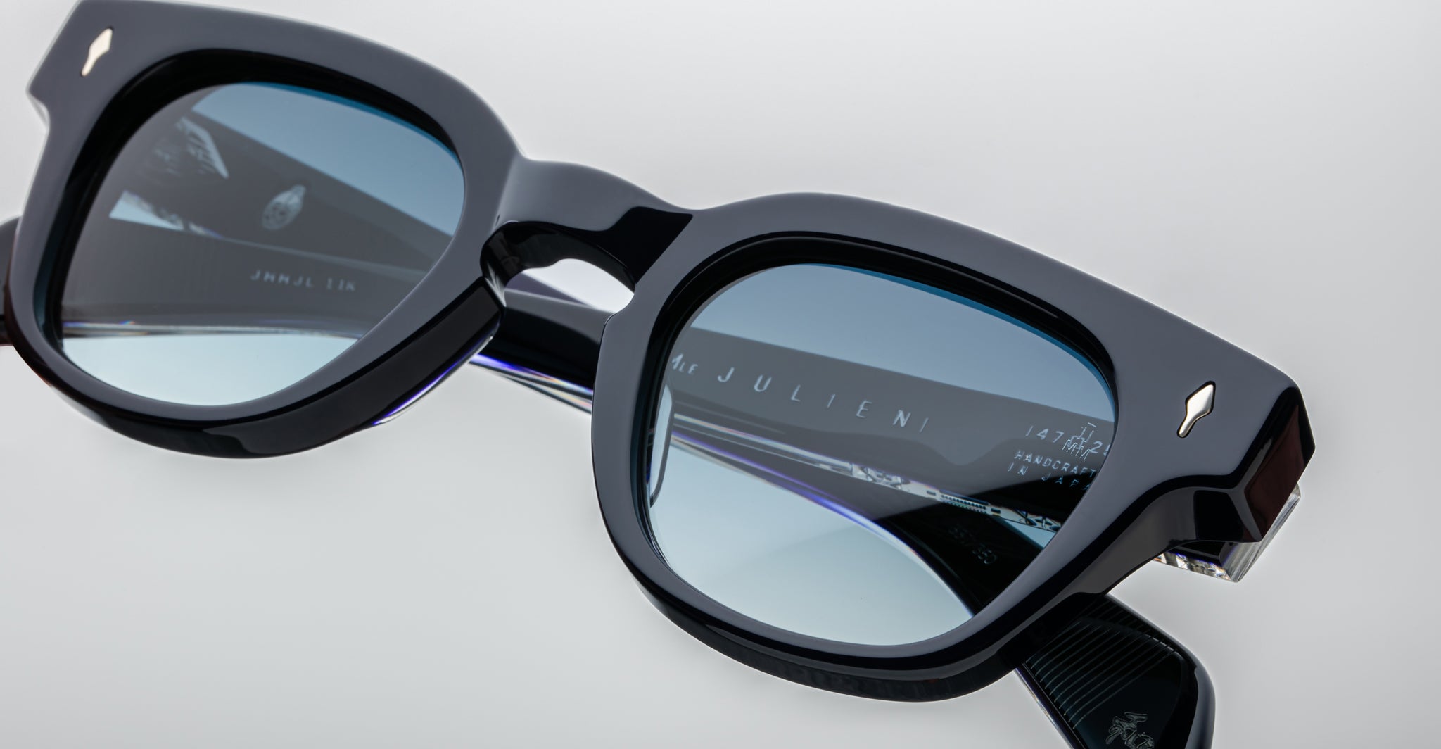 Close-up of black square-framed Jacques Marie Mage Julien sunglasses with blue-tinted lenses, displaying reflections and the engraved brand name on the inner side, set against a light background.