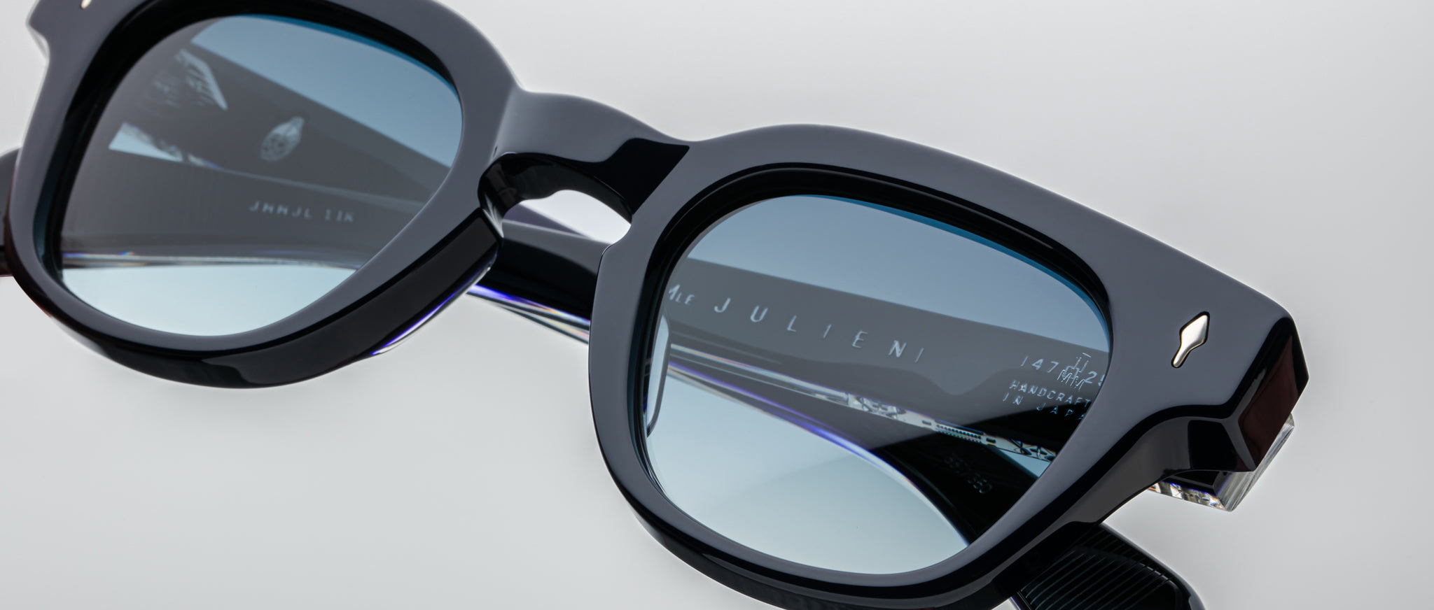 Close-up of black square-framed Jacques Marie Mage Julien sunglasses with blue-tinted lenses, displaying reflections and the engraved brand name on the inner side, set against a light background.