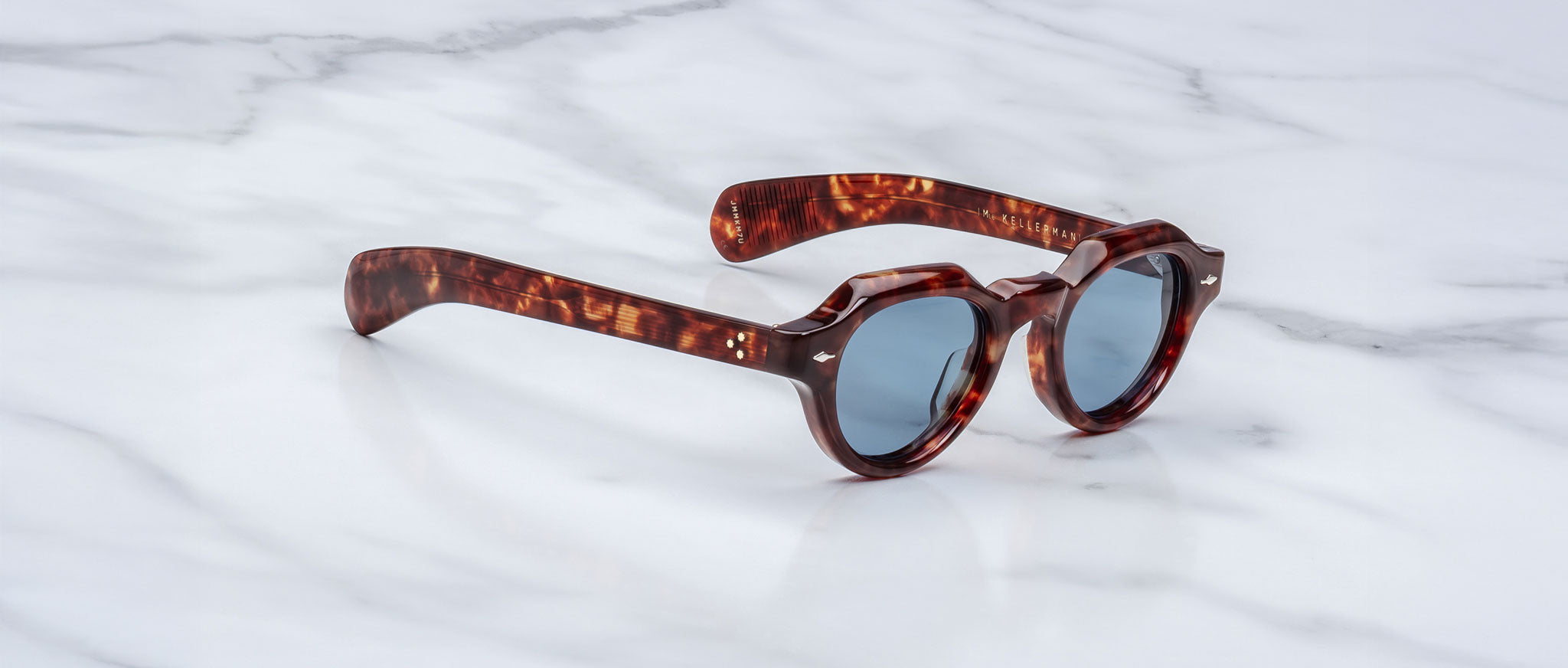 The Jacques Marie Mage Kellerman sunglasses, featuring round tortoiseshell frames and dark lenses, rest at an angle on a white marble surface, showcasing both their side profile and front view.
