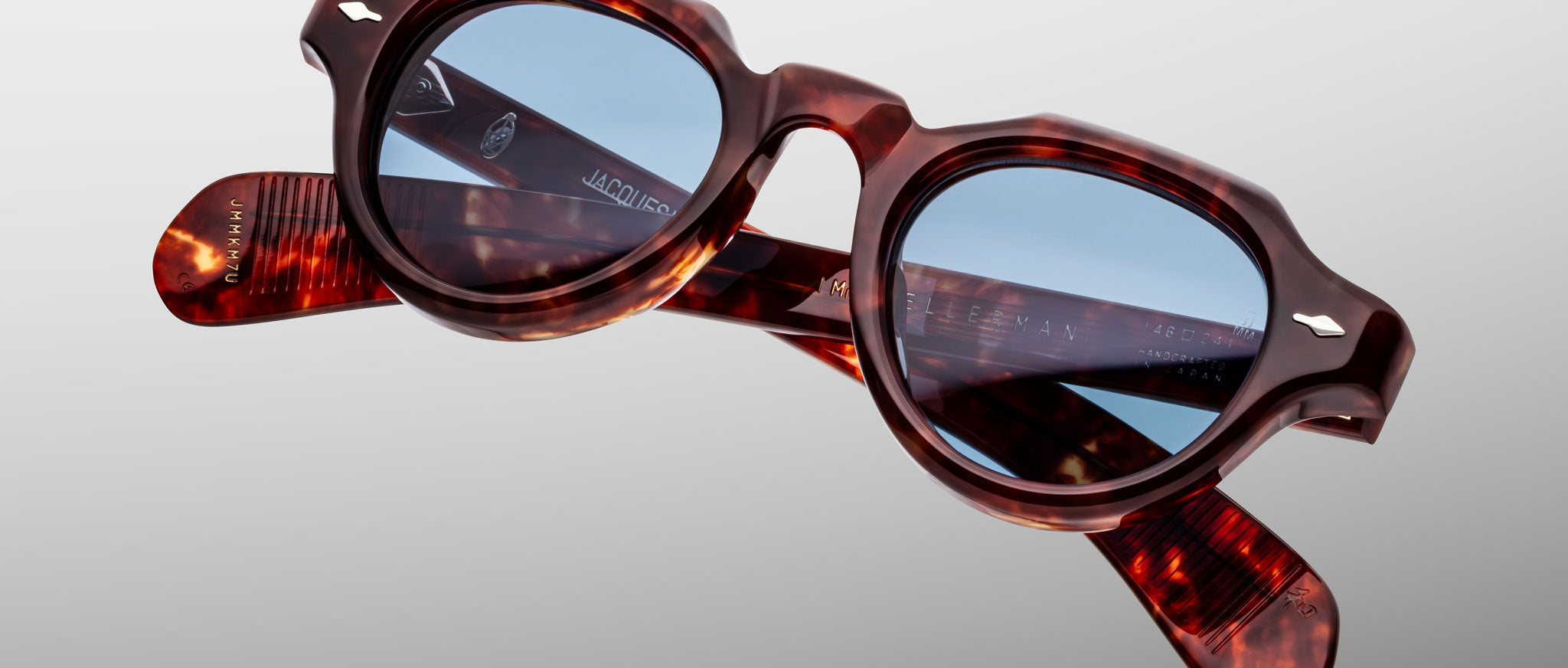 The Jacques Marie Mage Kellerman eyeglasses feature round, thick tortoiseshell frames with blue tinted lenses, shown against a plain light gray background with the glossy arms crossed.