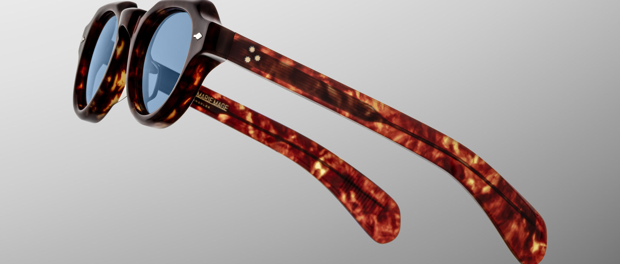 A pair of Jacques Marie Mage Kellerman tortoiseshell sunglasses with round blue lenses, shown at a three-quarter angle on a plain light gray background. The arms of the sunglasses are extended.
